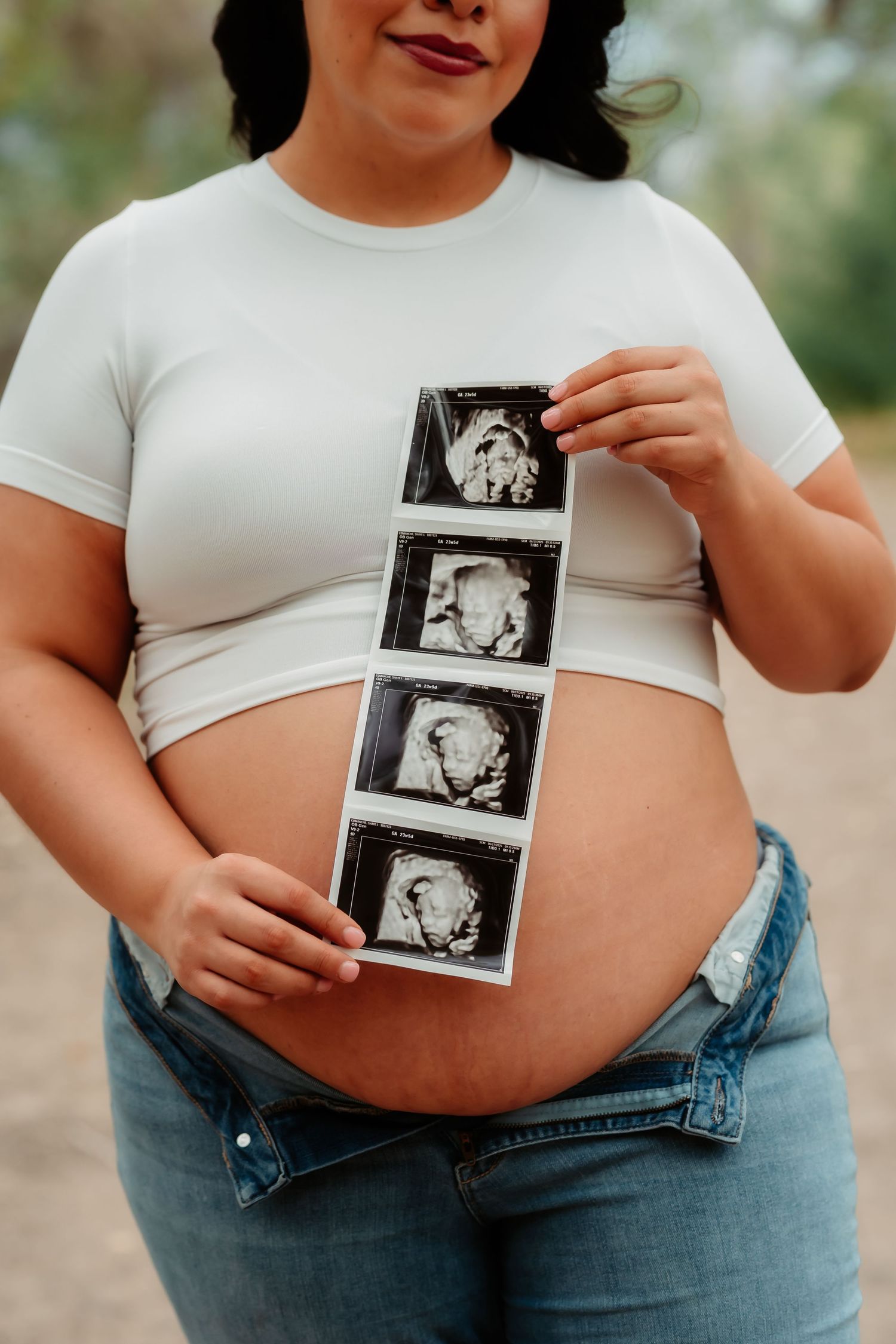 A series of ultrasound photos being held against a pregnant belly wearing jeans and a white top.