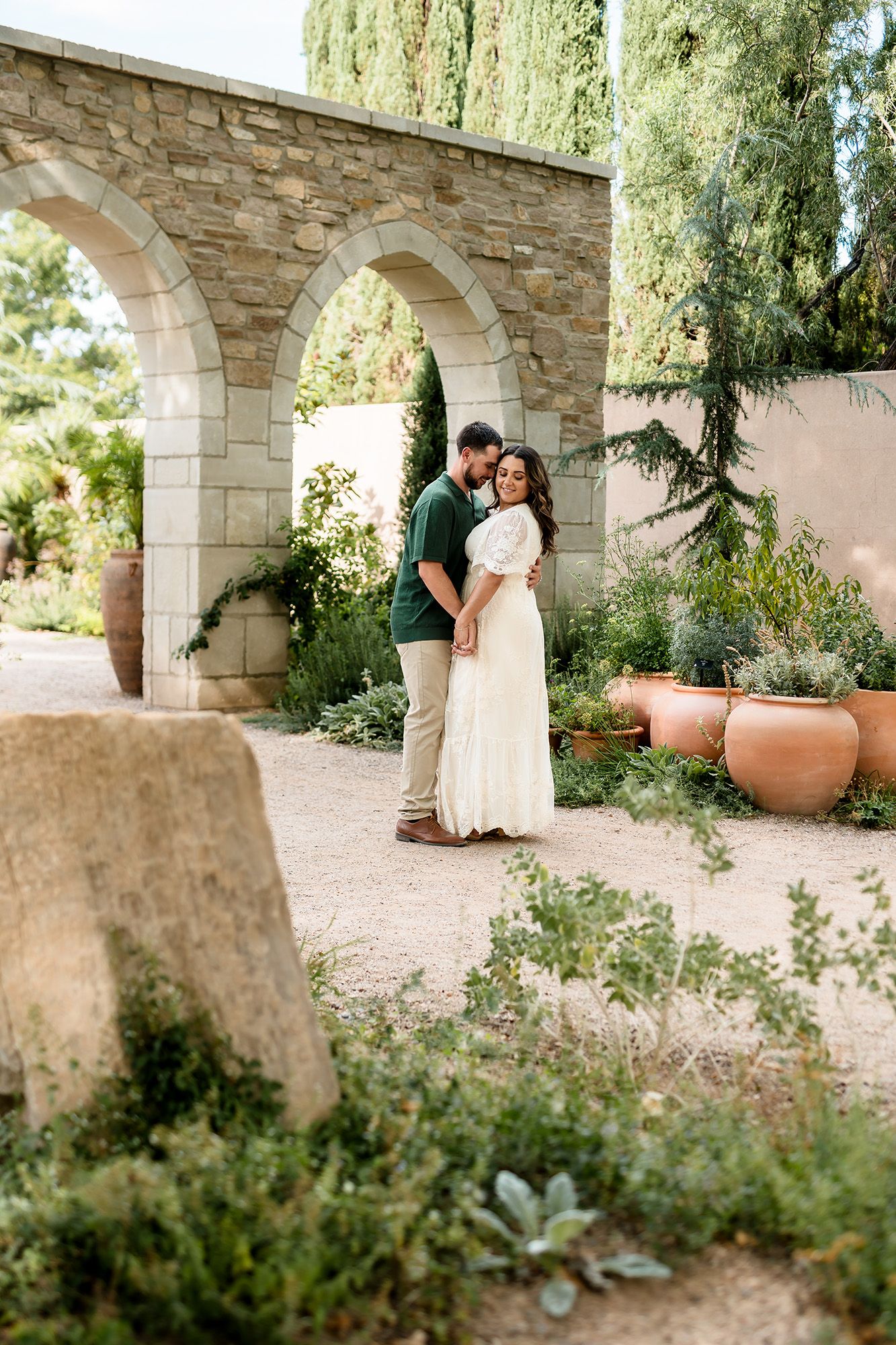 Couple embracing in a lush garden with stone arches, woman in white dress, man in green shirt.