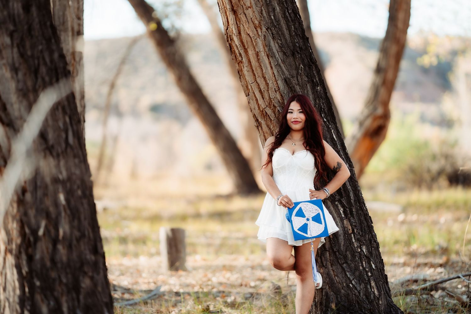 Graduate holding soccer ball poses near trees in outdoor setting.