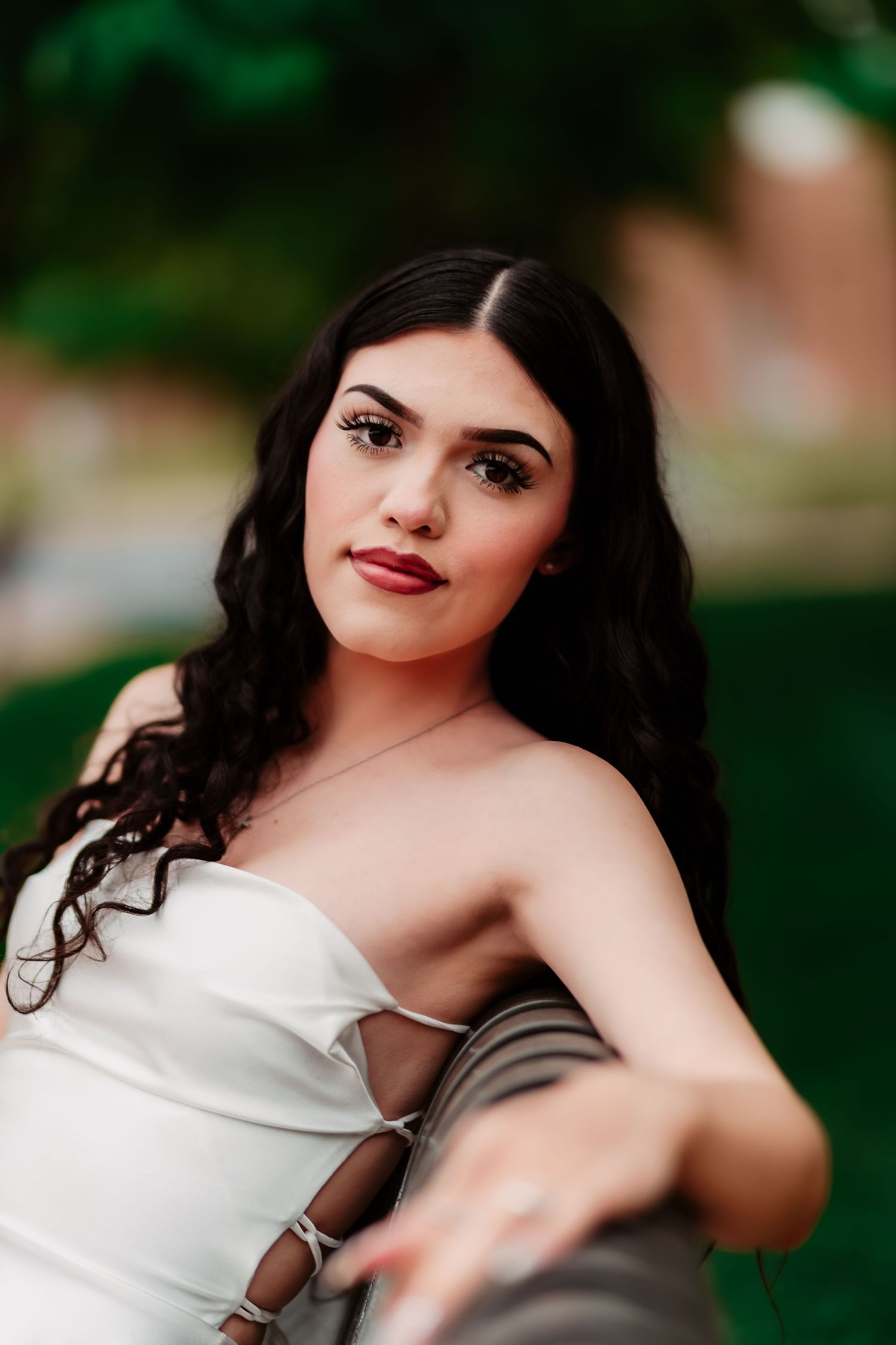 Portrait of someone in a white strapless dress with dark wavy hair against a blurred green background outdoors.