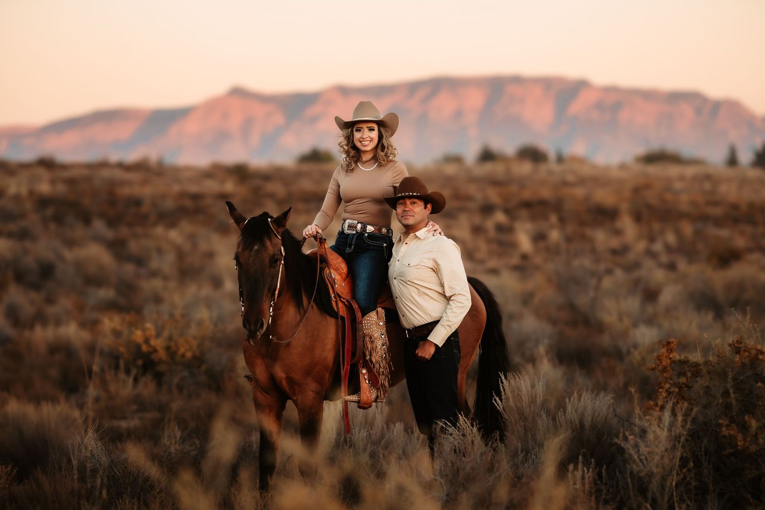 A series of horseback riders traverse a desert landscape during sunset with mountains in the background.