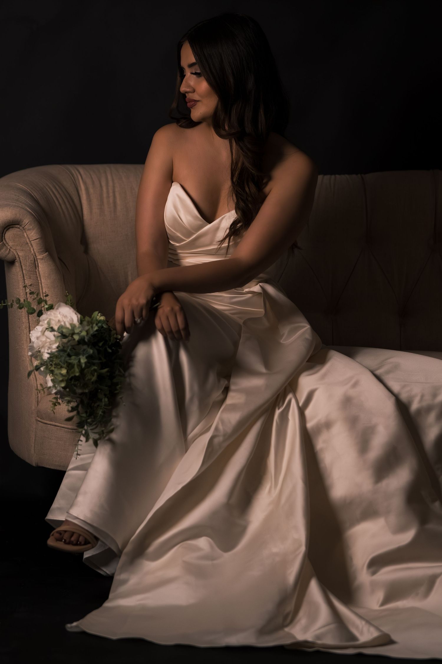 A bride in a white satin gown reclines on a couch holding a delicate floral bouquet in a moody dark setting.