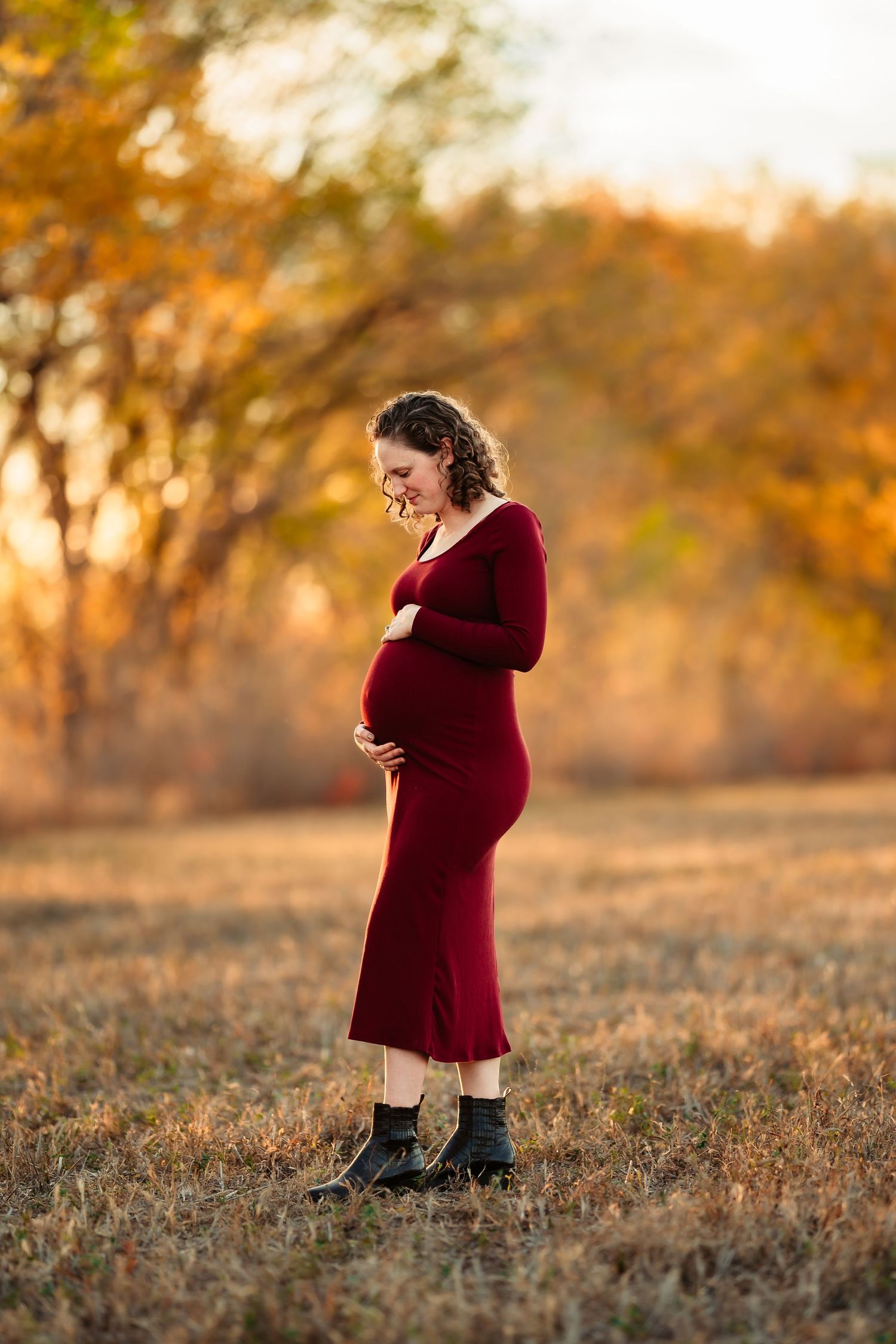 A person in a burgundy dress stands in a golden autumn field during sunset, creating a warm silhouetted effect.