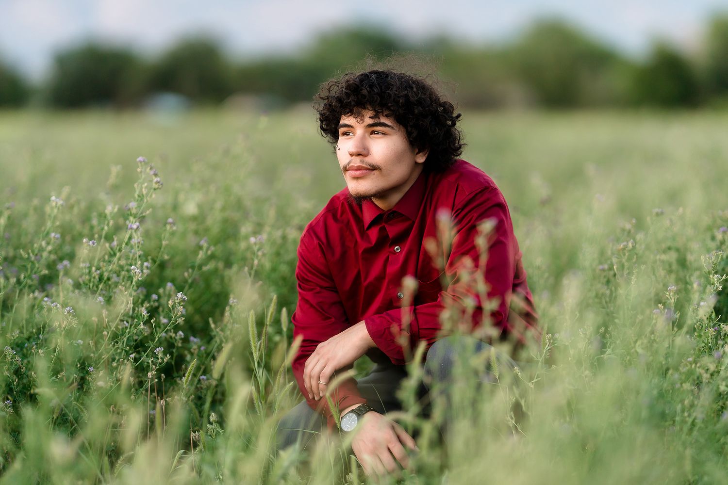 Young man with curly hair in red shirt sitting in lush green field, gazing thoughtfully into the distance.