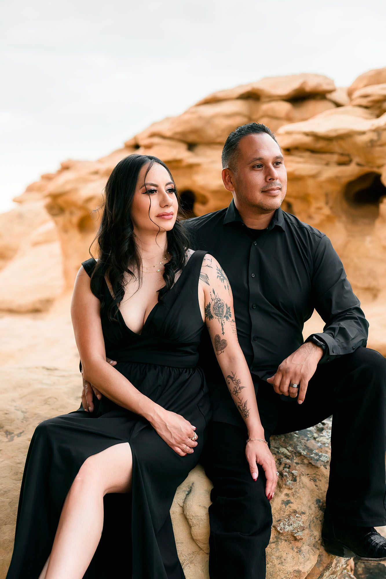 Couple in matching black outfits sitting on desert rocks, woman in black gown with tattoos.
