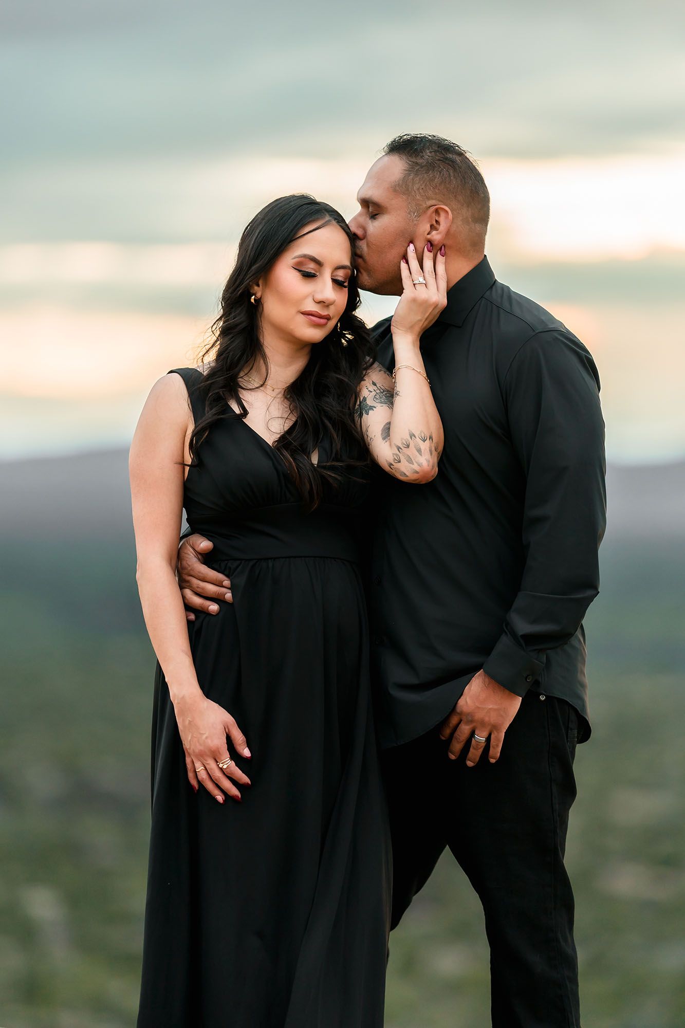 Pregnant woman in black gown kissed on cheek by tattooed man in black shirt at sunset.