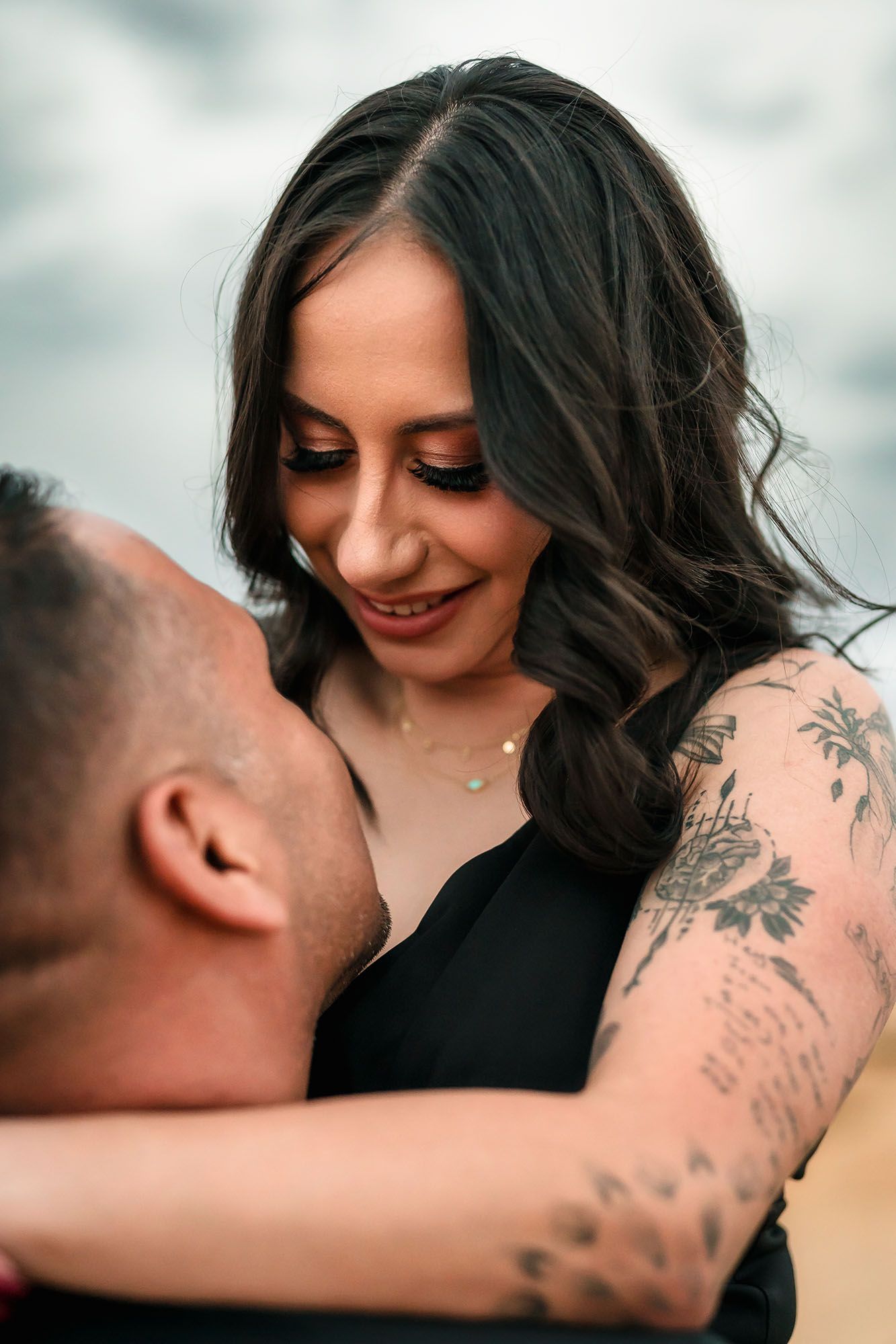 Close-up of smiling brunette woman with tattoos embracing her partner under cloudy skies.