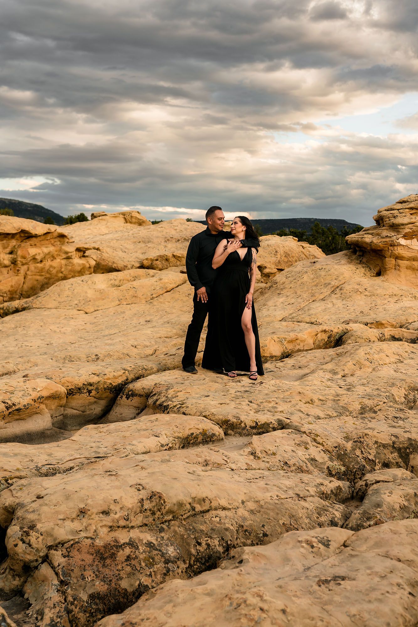 Romantic couple in black formal wear embracing on golden sandstone rocks under dramatic cloudy sky.