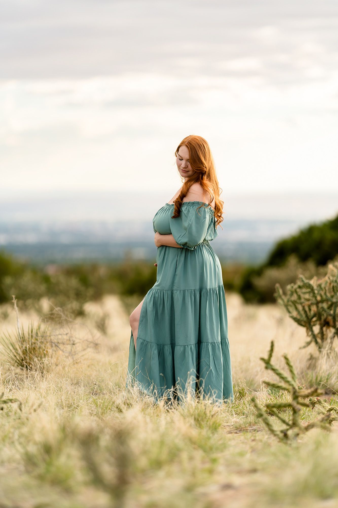A red-haired woman in a teal off-shoulder maxi dress stands gracefully in a desert field under a cloudy sky.