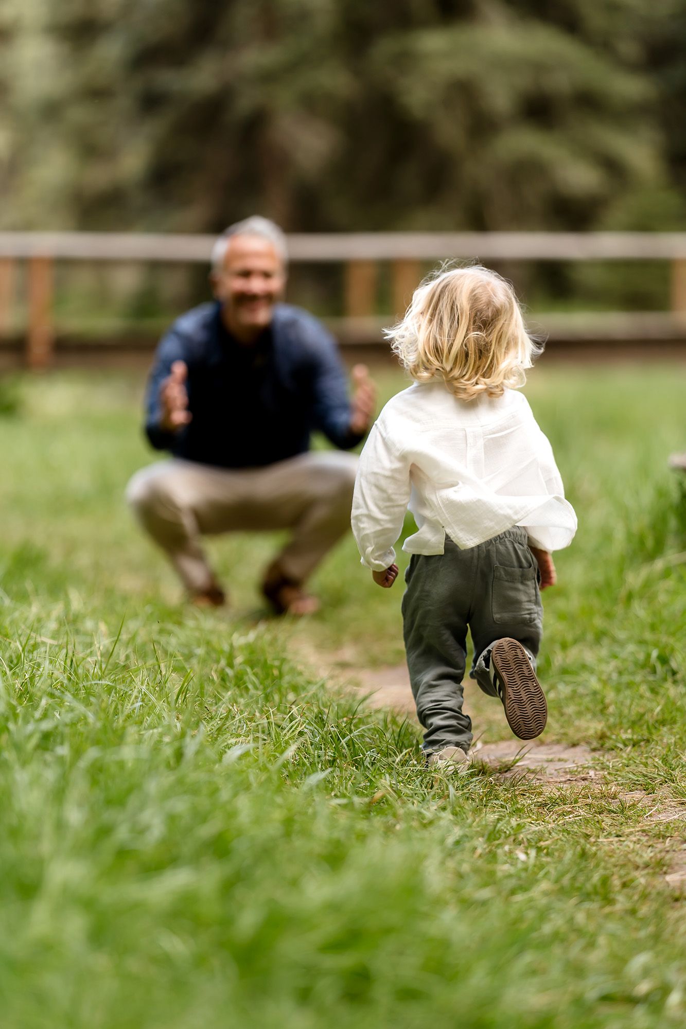 A young child with blonde hair runs toward a smiling man crouching with open arms on a lush green garden path.