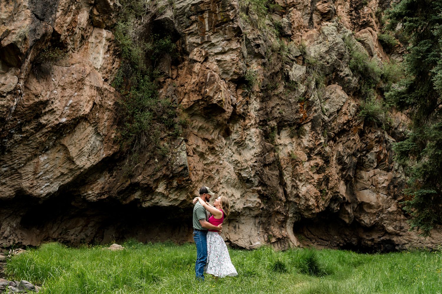 Couple embracing in front of a dramatic rocky cliff face surrounded by lush green grass in a mountain setting.
