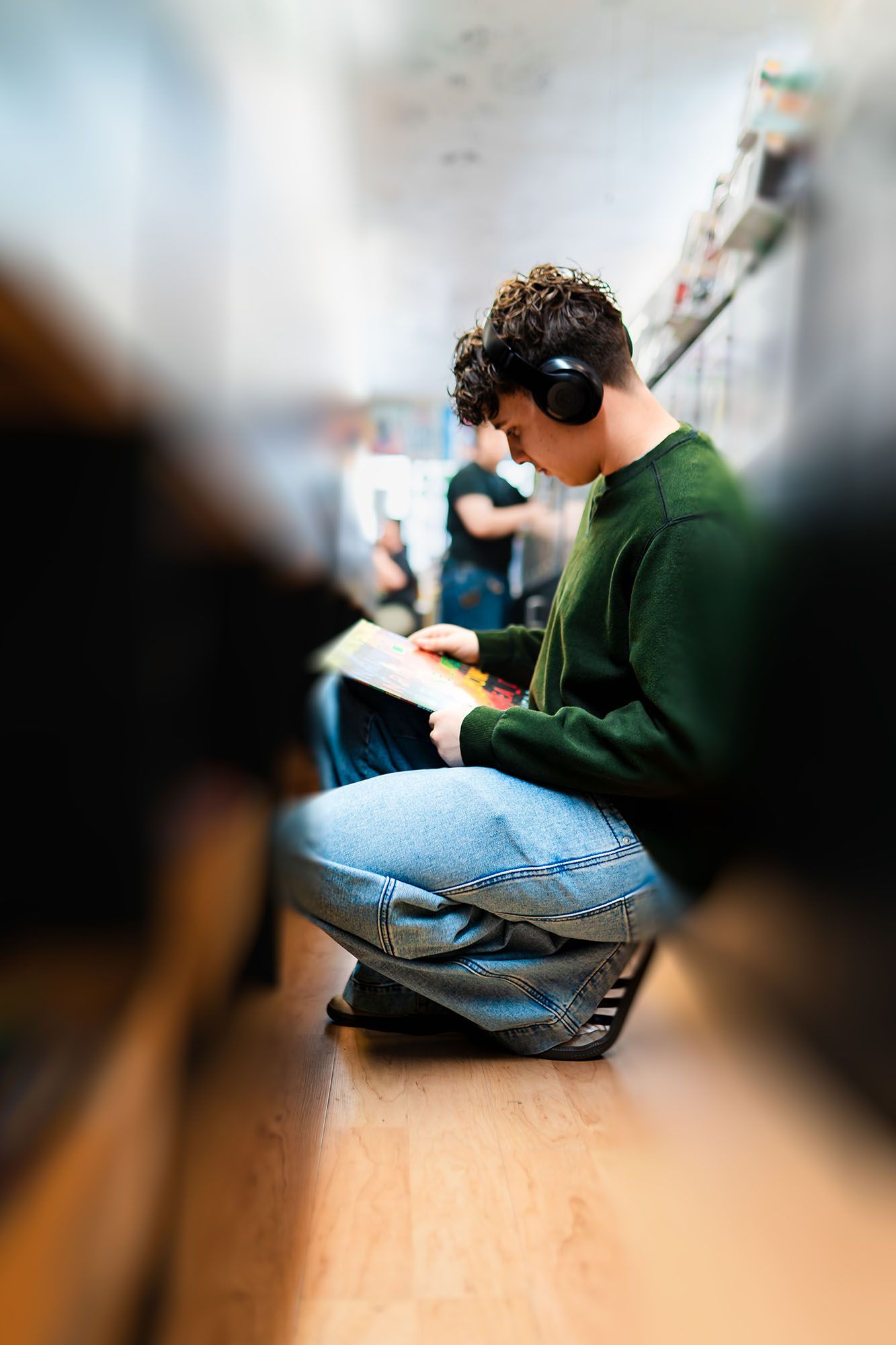 Curly-haired teen wearing headphones reading a book while sitting on hallway floor at school.