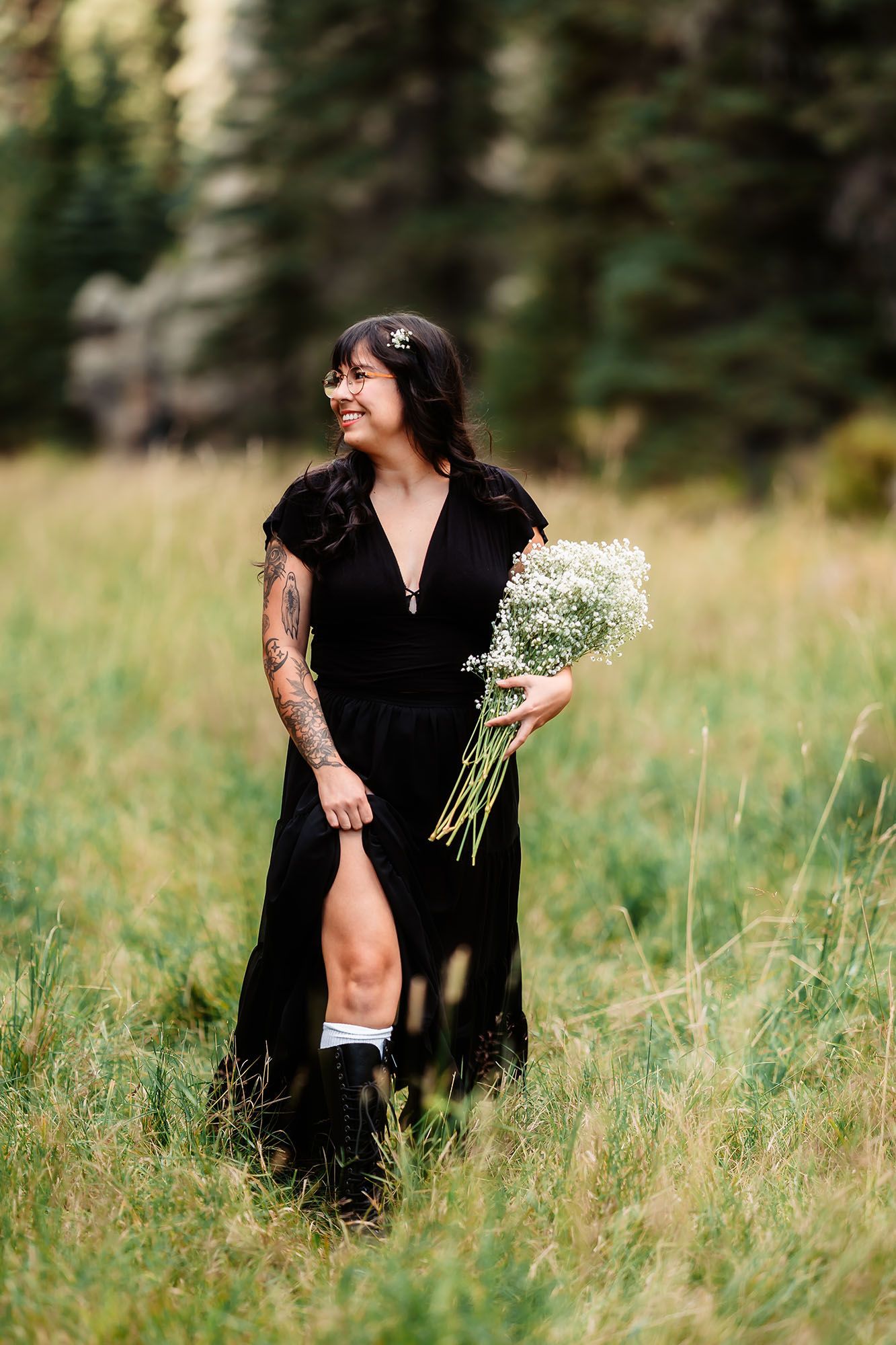 Woman in black dress holds white wildflowers while standing in a lush green meadow.