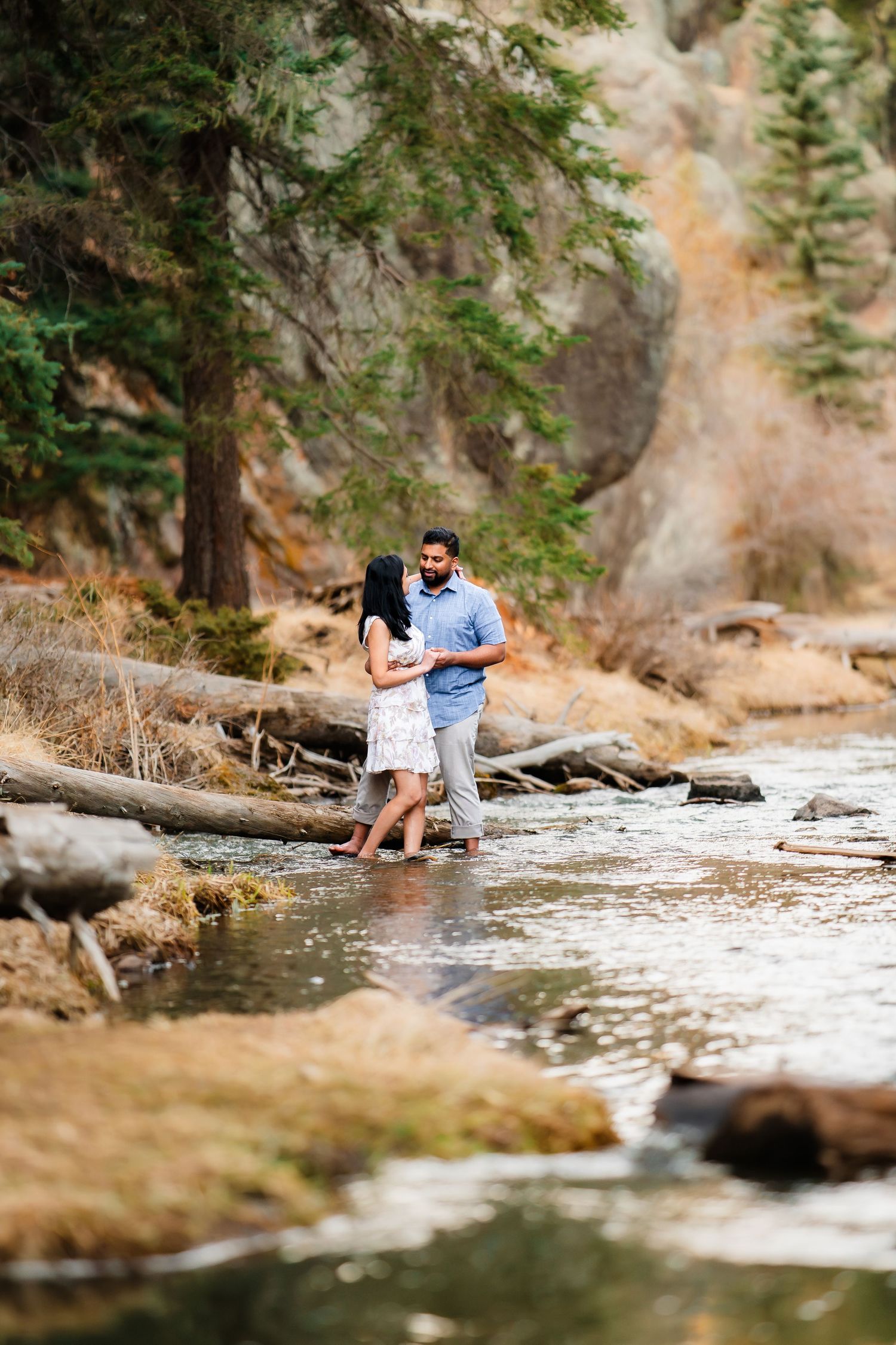 Two figures walk along a scenic riverside trail surrounded by evergreen trees and natural rock formations.