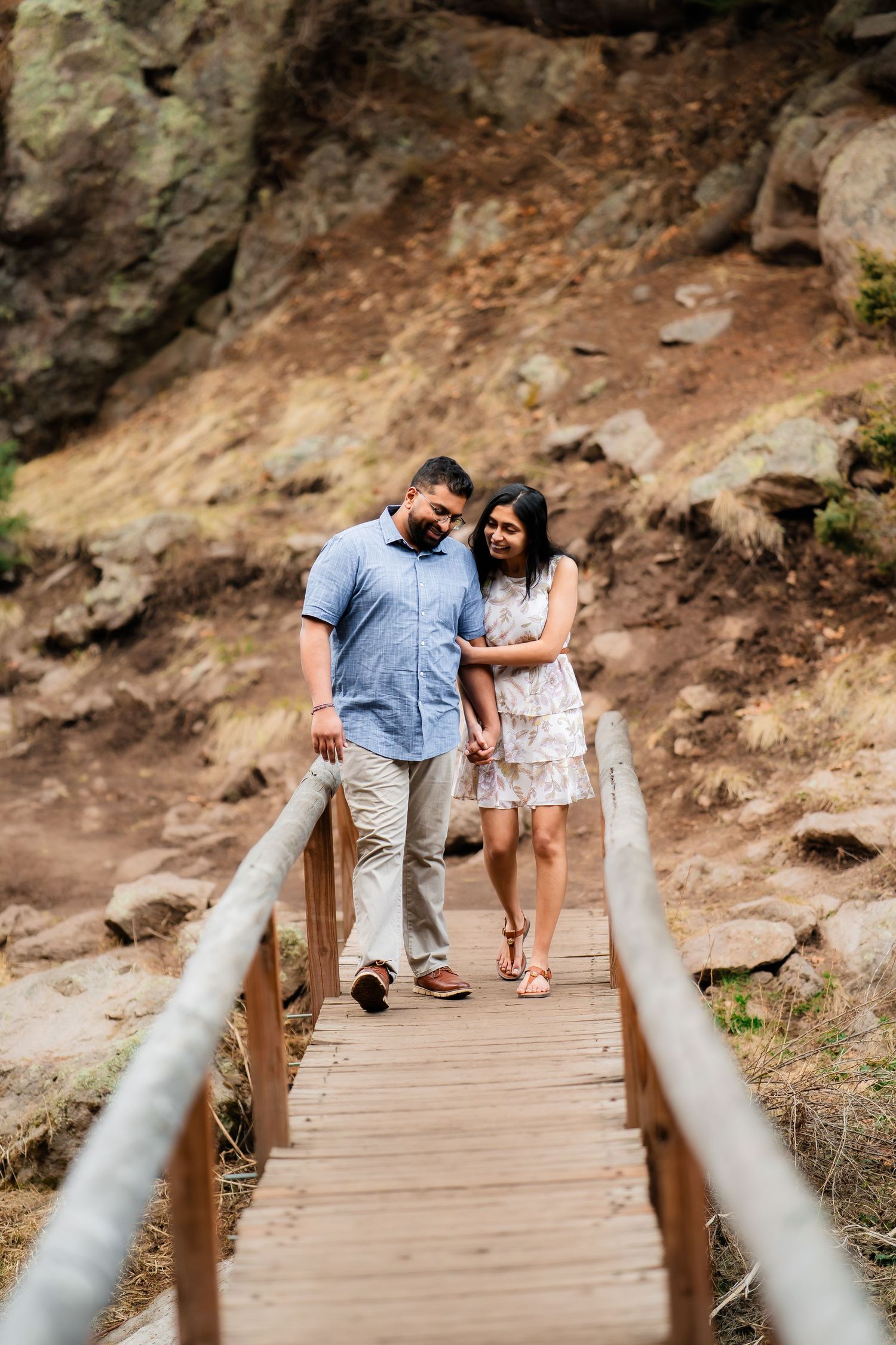 Couple walks together holding hands on wooden boardwalk trail through rocky terrain.