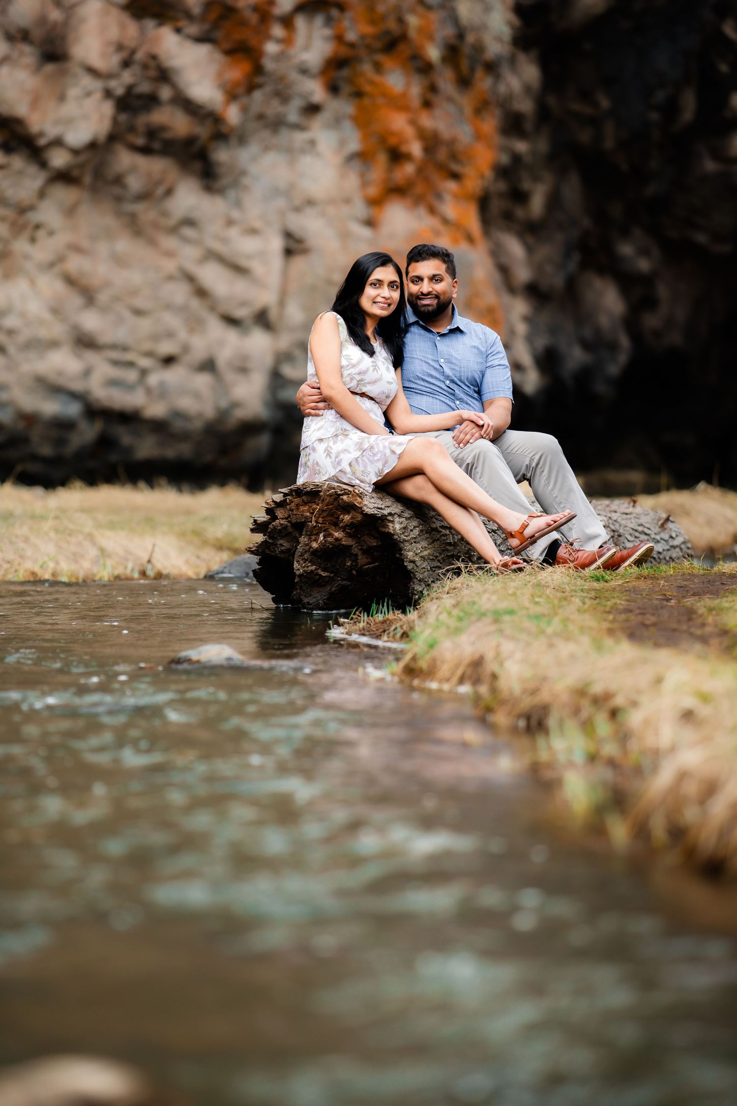 A couple sits together by a stream with rocky cliffs in the background.