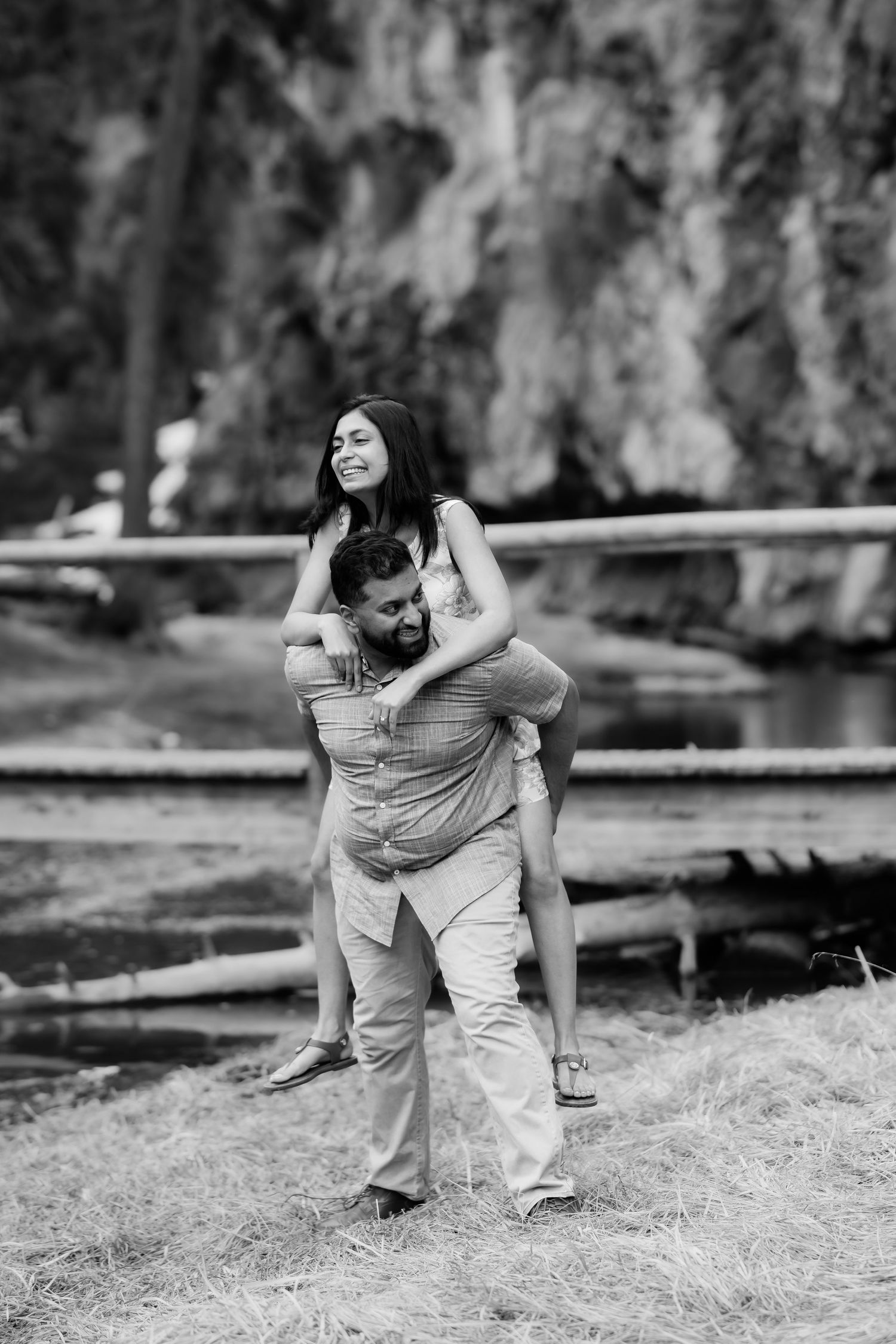 A couple enjoys a playful piggyback ride along a scenic riverbank in a black and white photo.