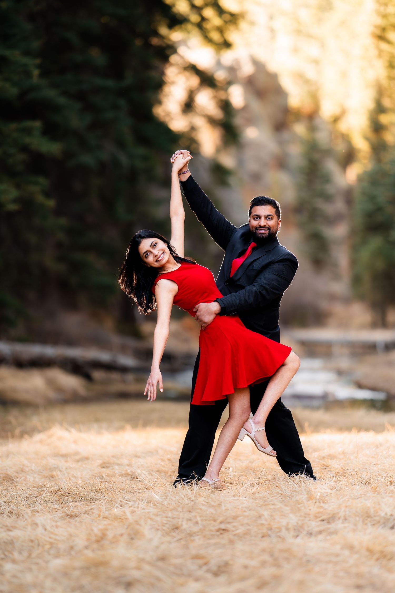 A couple performs an elegant dance pose outdoors with the woman in a flowing red dress against a natural backdrop.