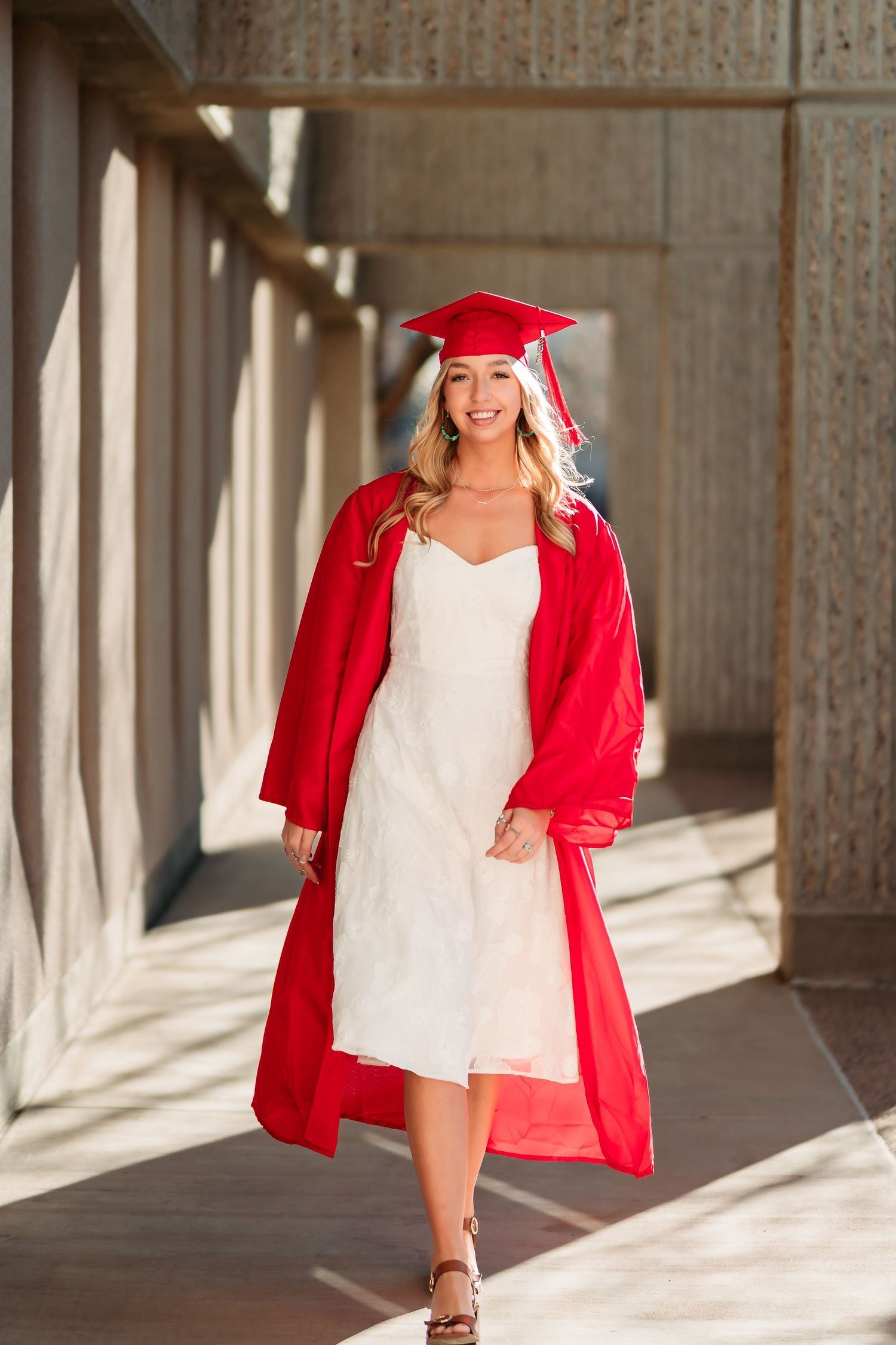 Graduate in red cap and gown with white dress walks through sunlit concrete hallway during graduation photoshoot.