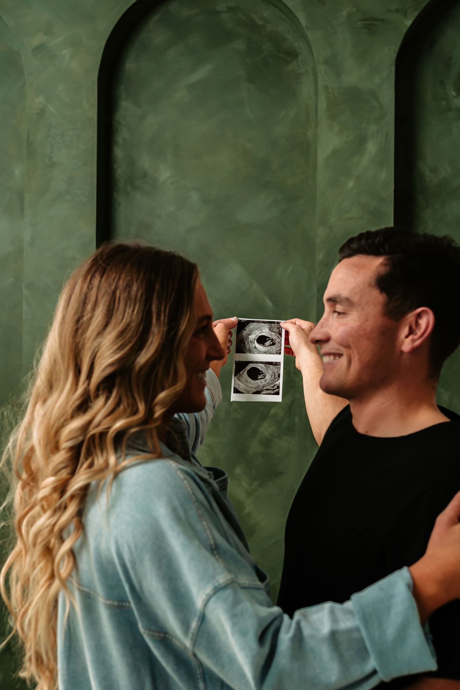 Couple playfully poses with ultrasound photo against green textured wall.