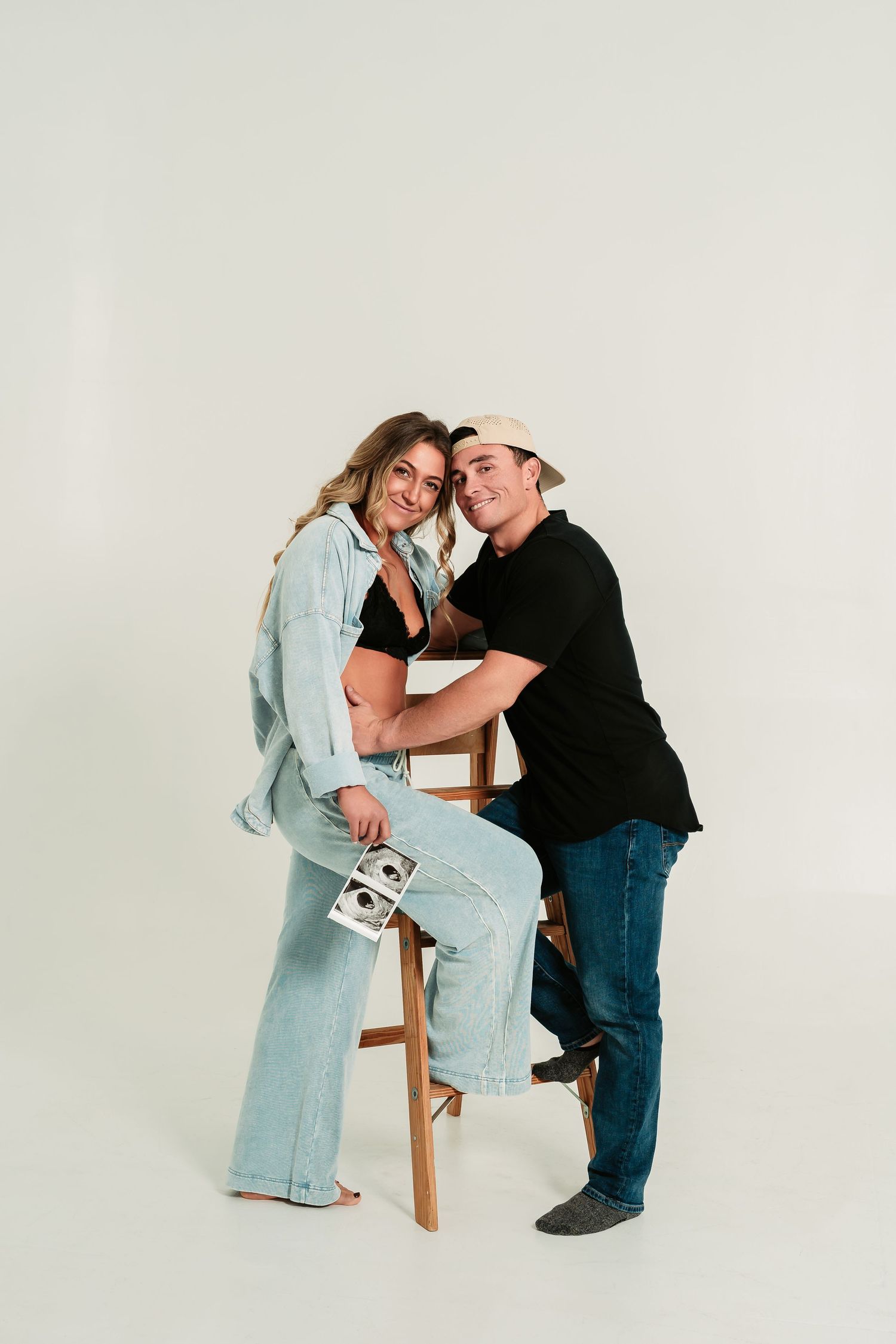 A couple in casual attire shares an intimate moment near a wooden stool against a white backdrop.