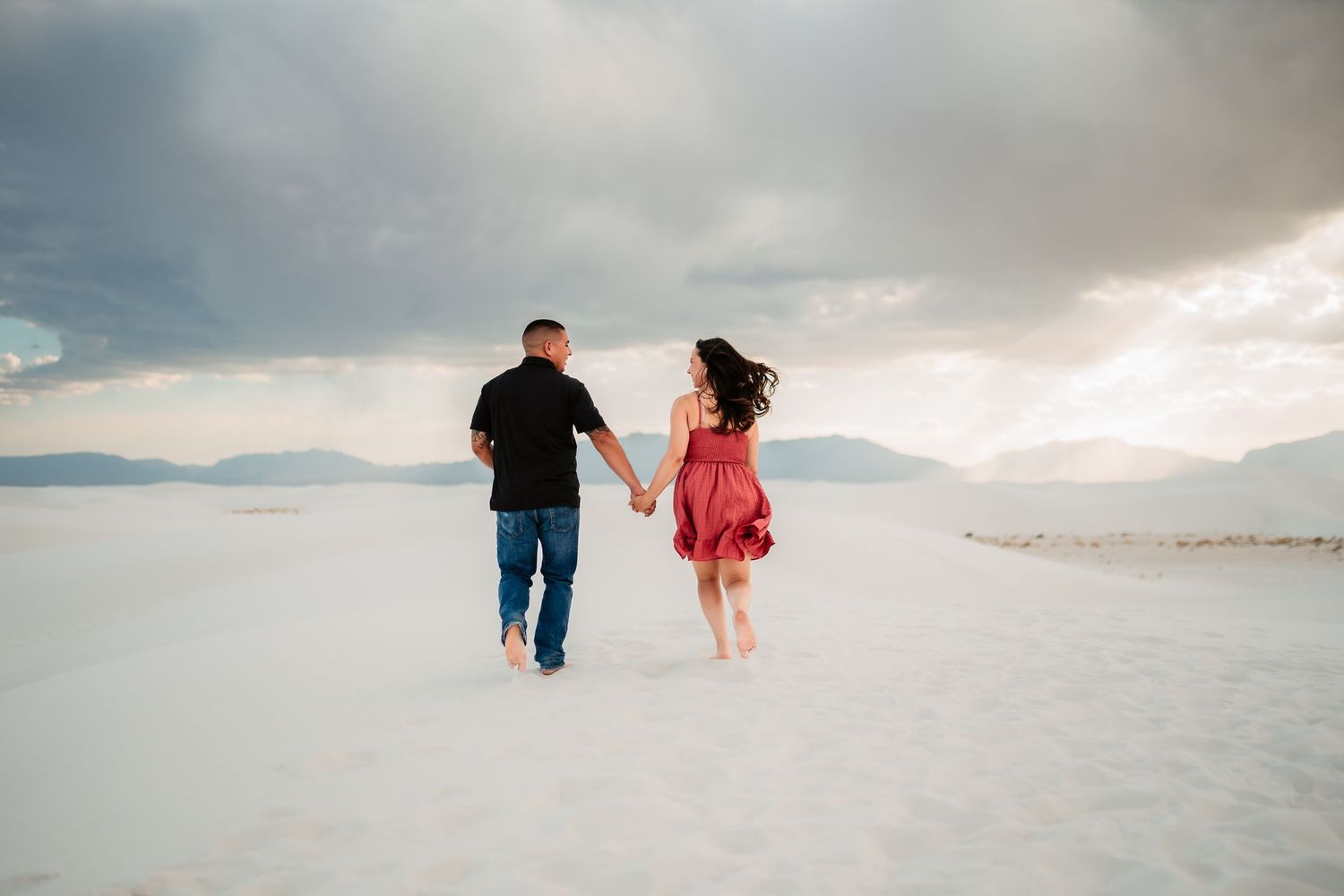 A couple holding hands while walking on the white sands of White Sands National Park with dramatic clouds overhead.