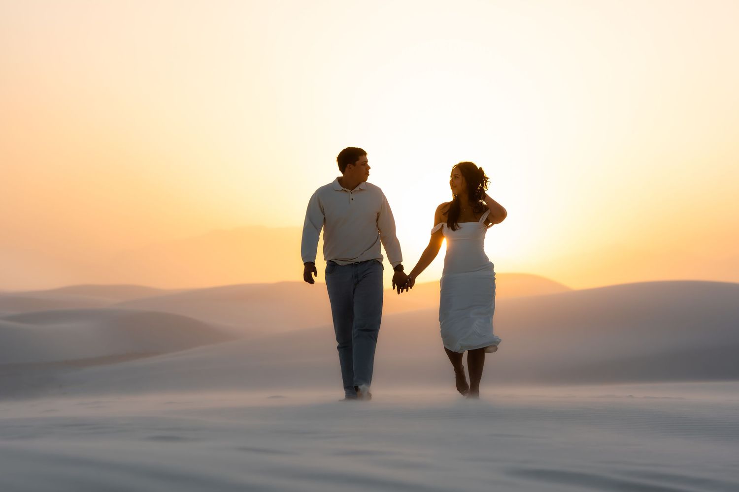 A couple walks hand in hand on sandy dunes during a golden sunset creating silhouettes against the warm sky.