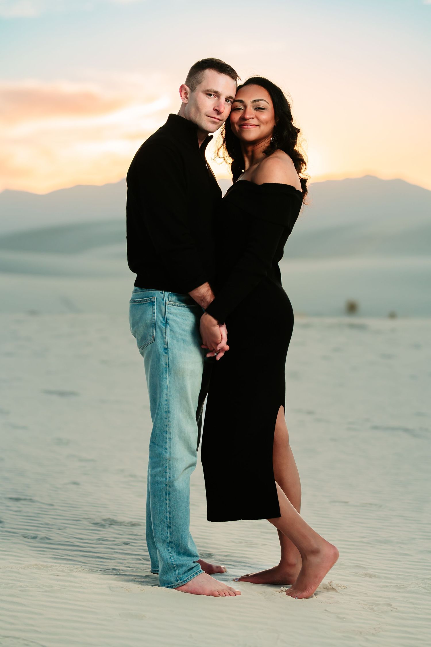 A romantic couple embraces on white sand dunes at sunset with mountains in the background.
