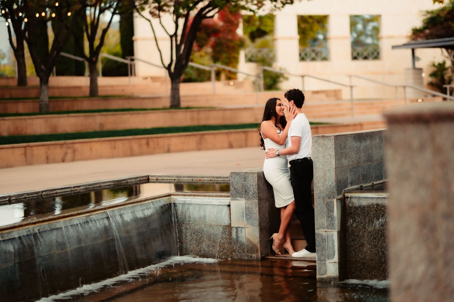 Two people share an intimate moment near a stone wall with soft lighting and architectural details.