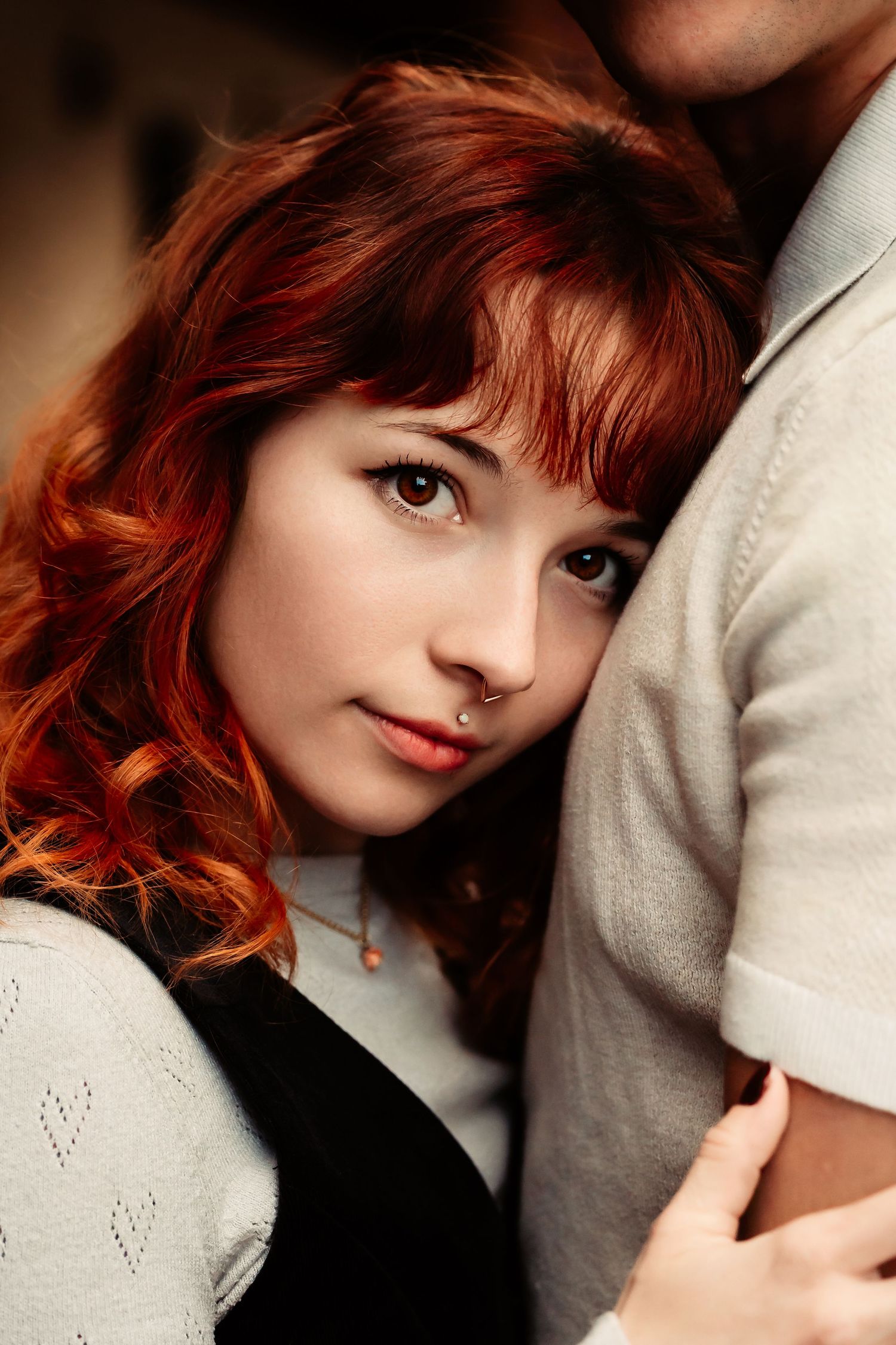 A woman with vibrant red hair and bangs poses intimately against someone wearing a light colored sweater.