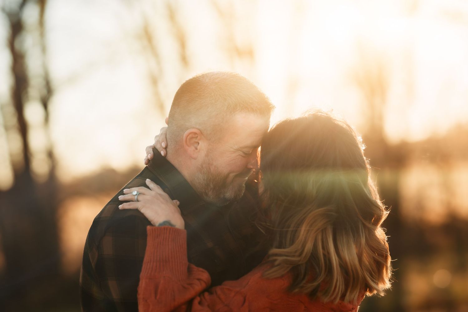Couple embraces in warm sunset light with sun flare and trees silhouetted in background.