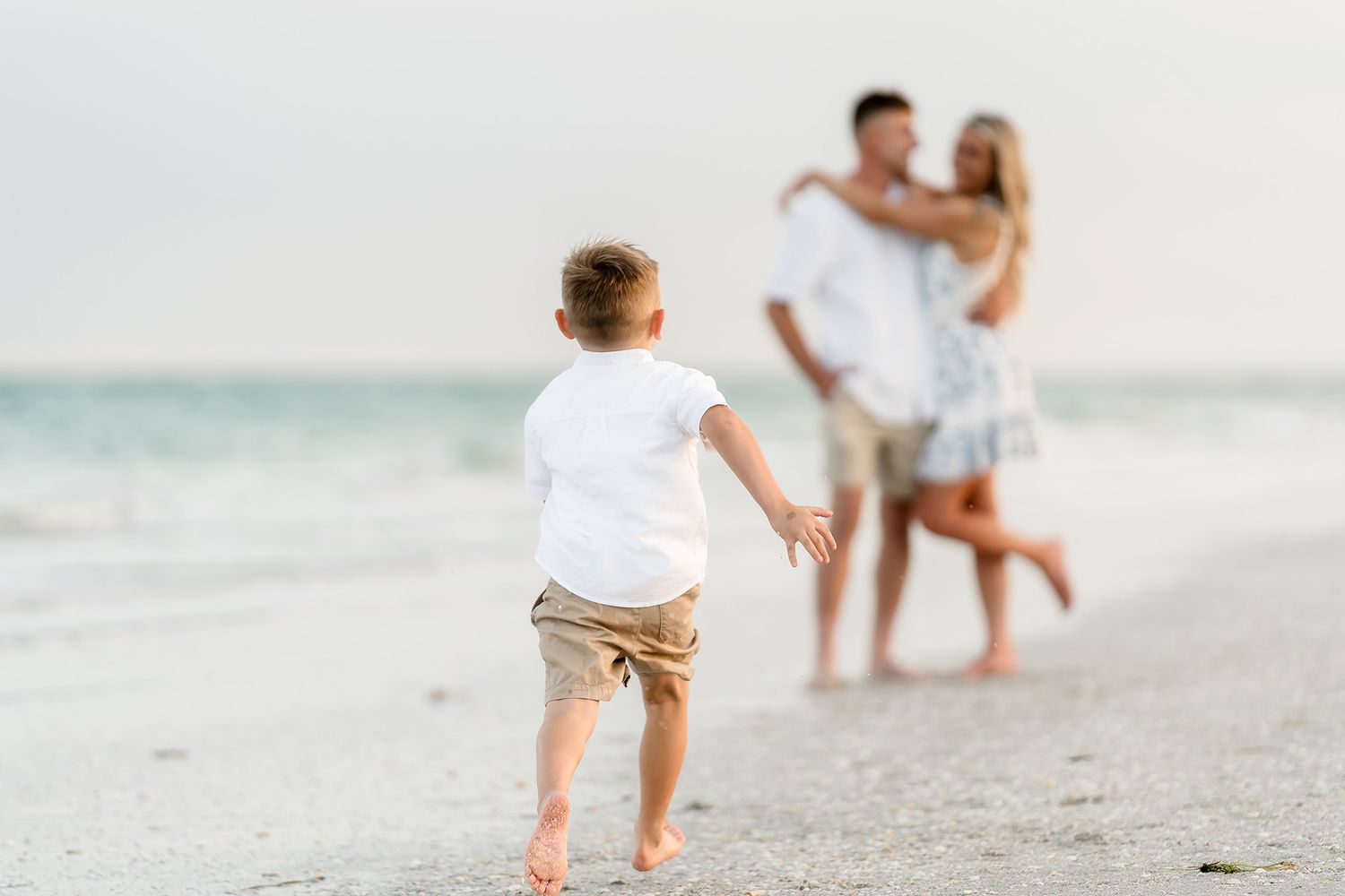 Young couple embraces on sandy beach as toddler boy runs toward them at sunset.