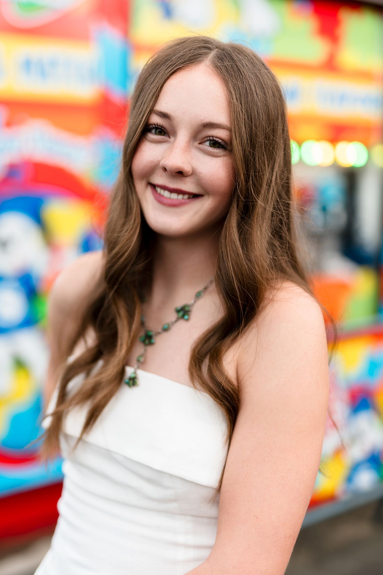 Young woman in white strapless dress with green necklace smiling in front of colorful background.