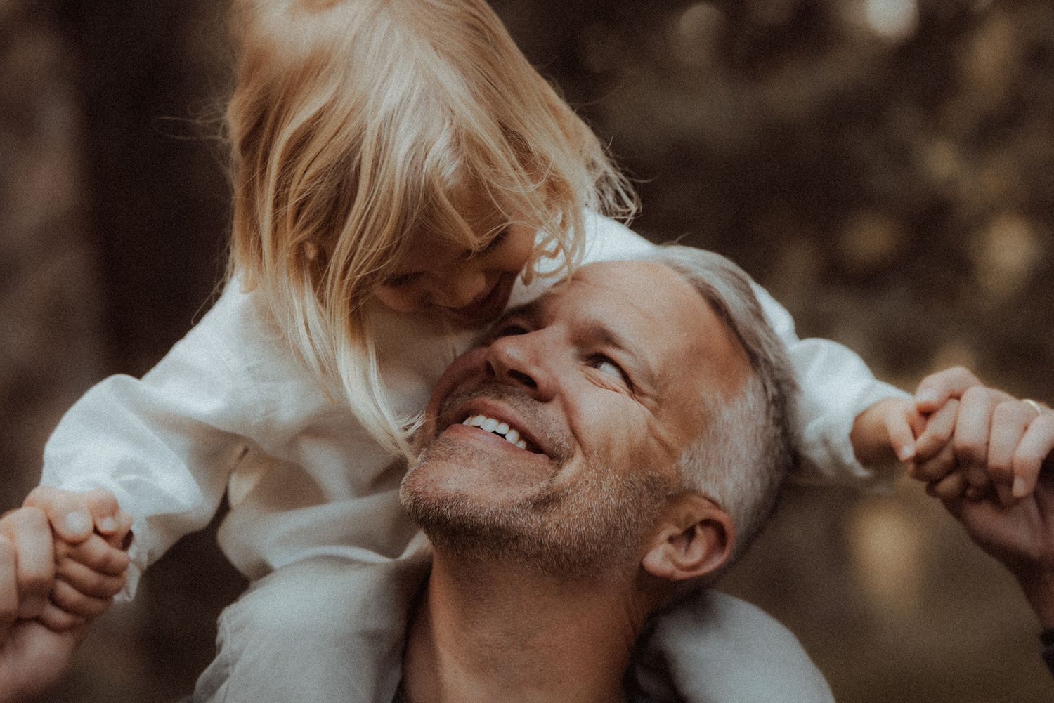 Smiling father carrying young blonde girl on his shoulders in a sunlit forest.