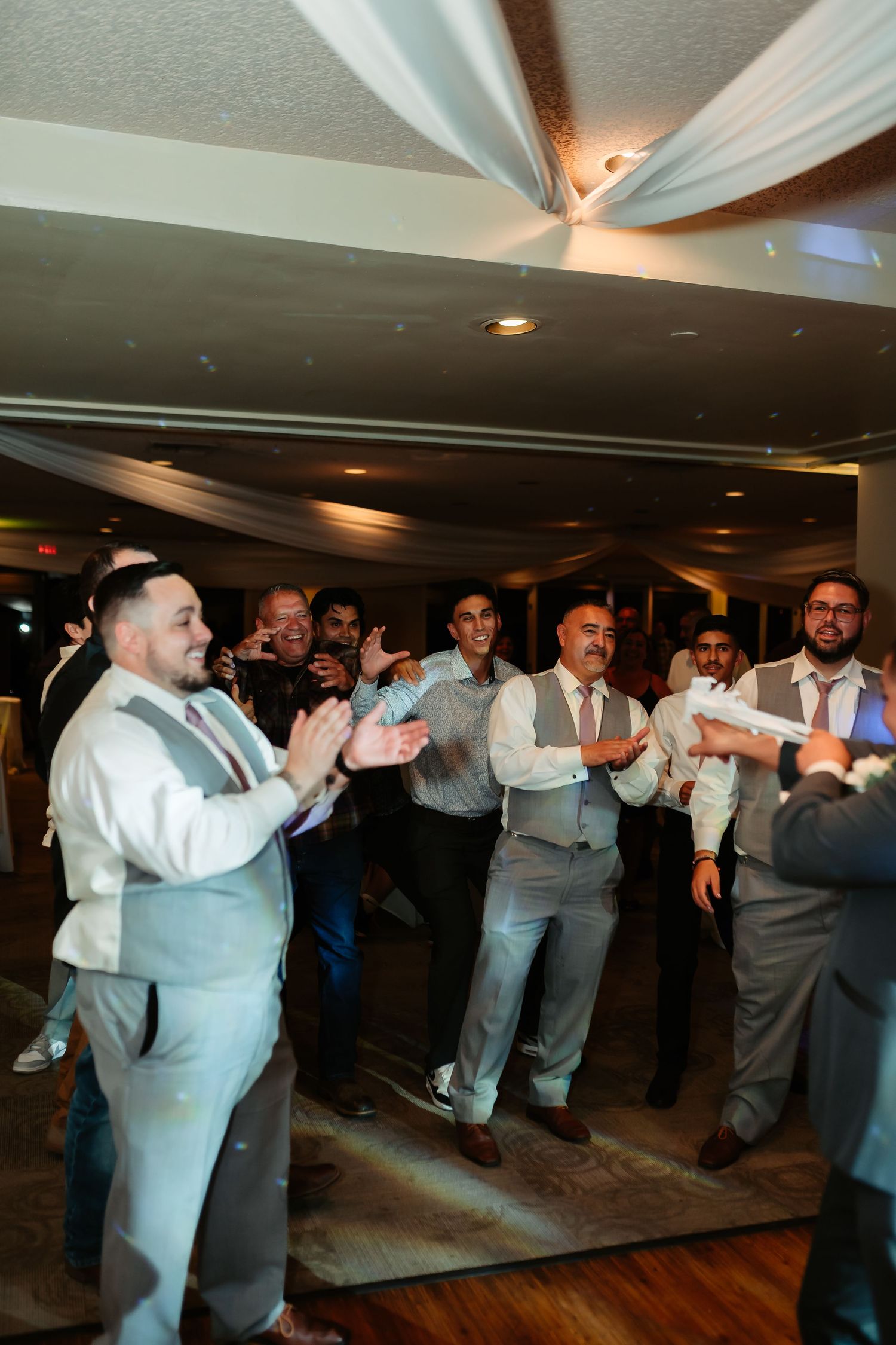 Groomsmen in formal attire performing a synchronized dance line at a wedding reception on a wooden dance floor.