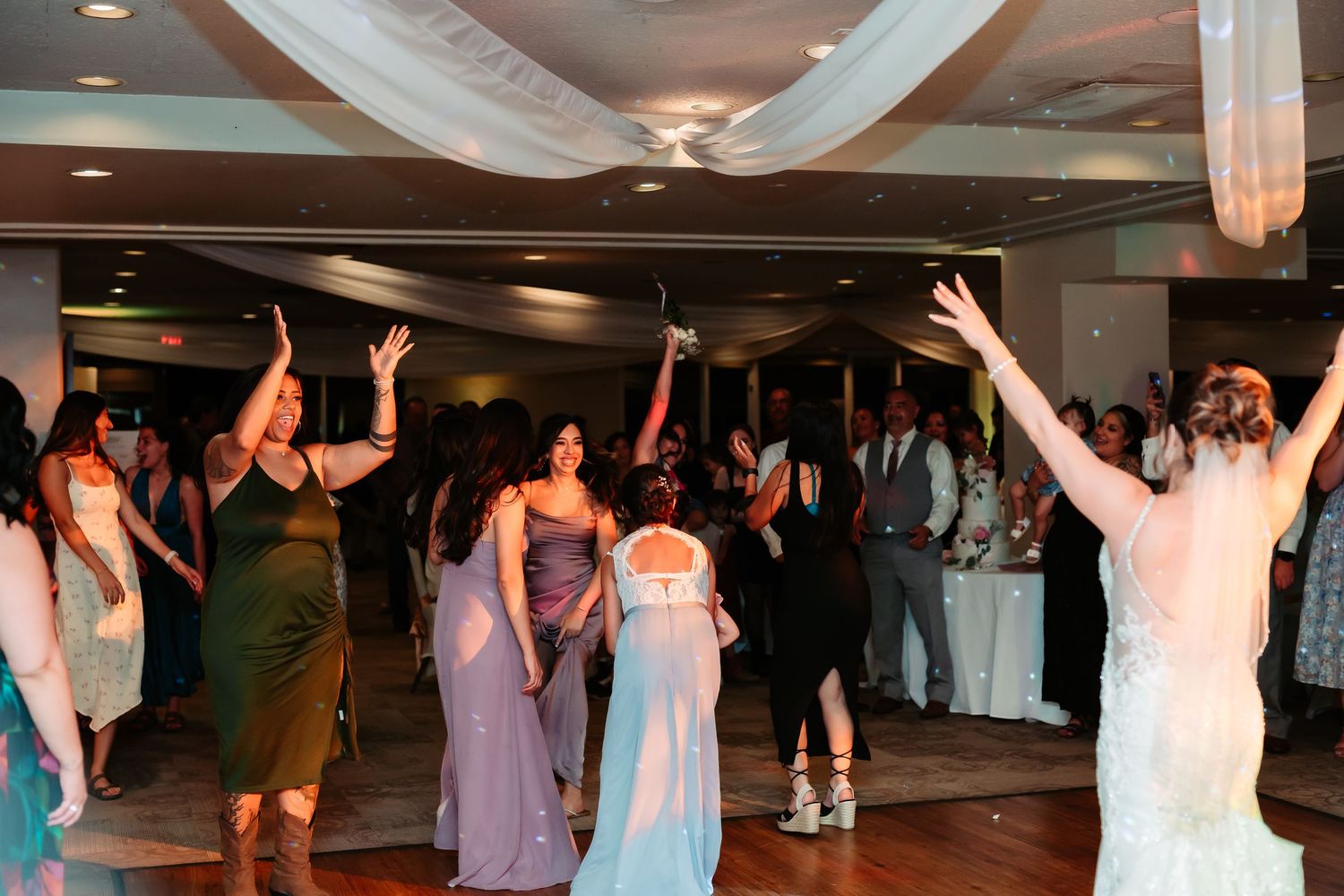 Wedding guests dancing and celebrating on a crowded dance floor with white ceiling drapes.