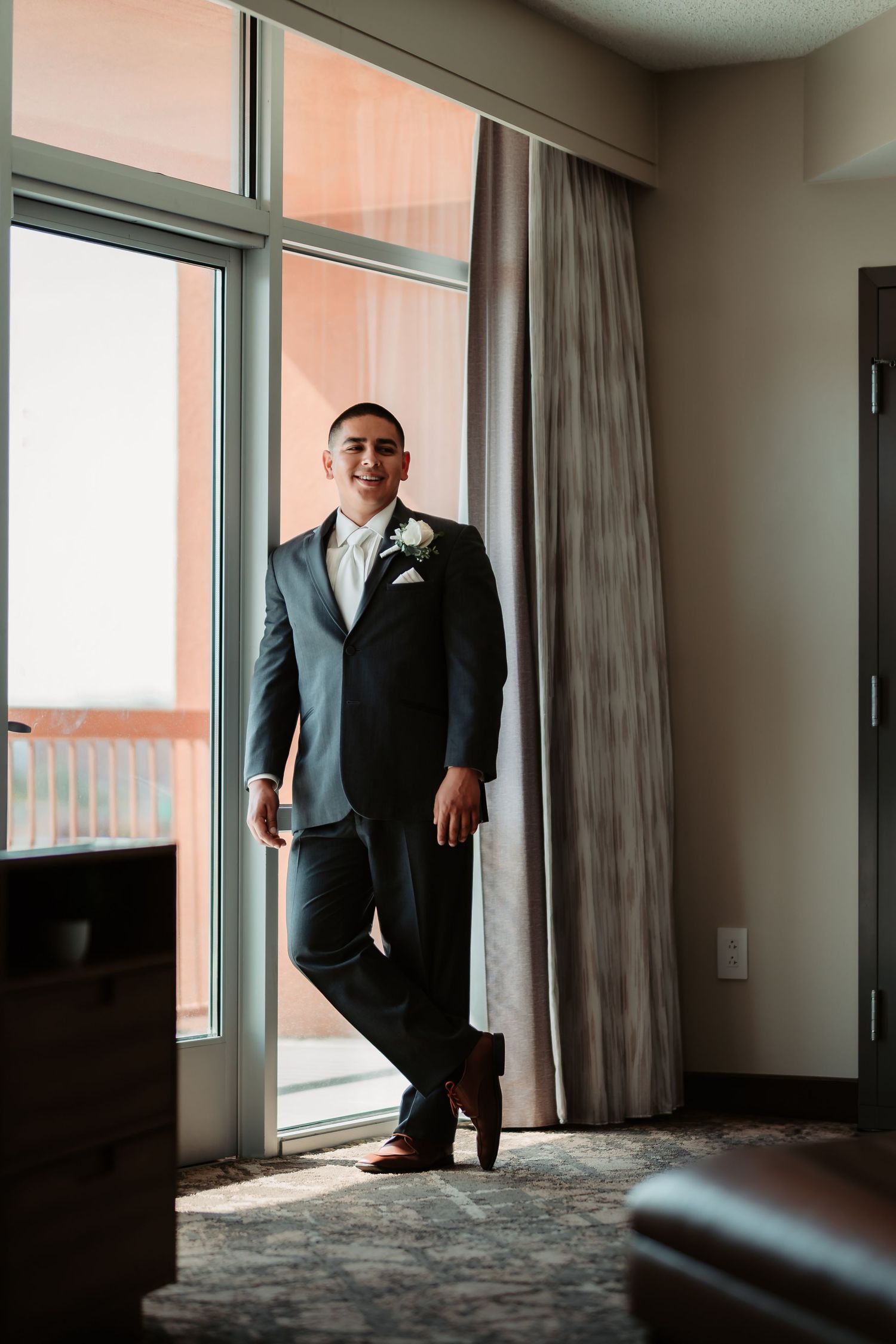 Man in formal dark suit stands by large window in elegant hotel room with natural lighting and grey carpeting.