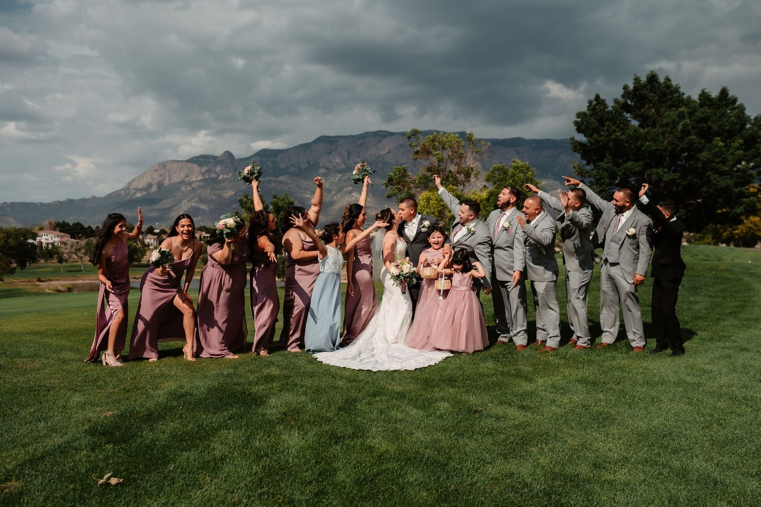 Wedding party lies on grassy lawn with mountains and cloudy sky in panoramic collage of celebratory poses.