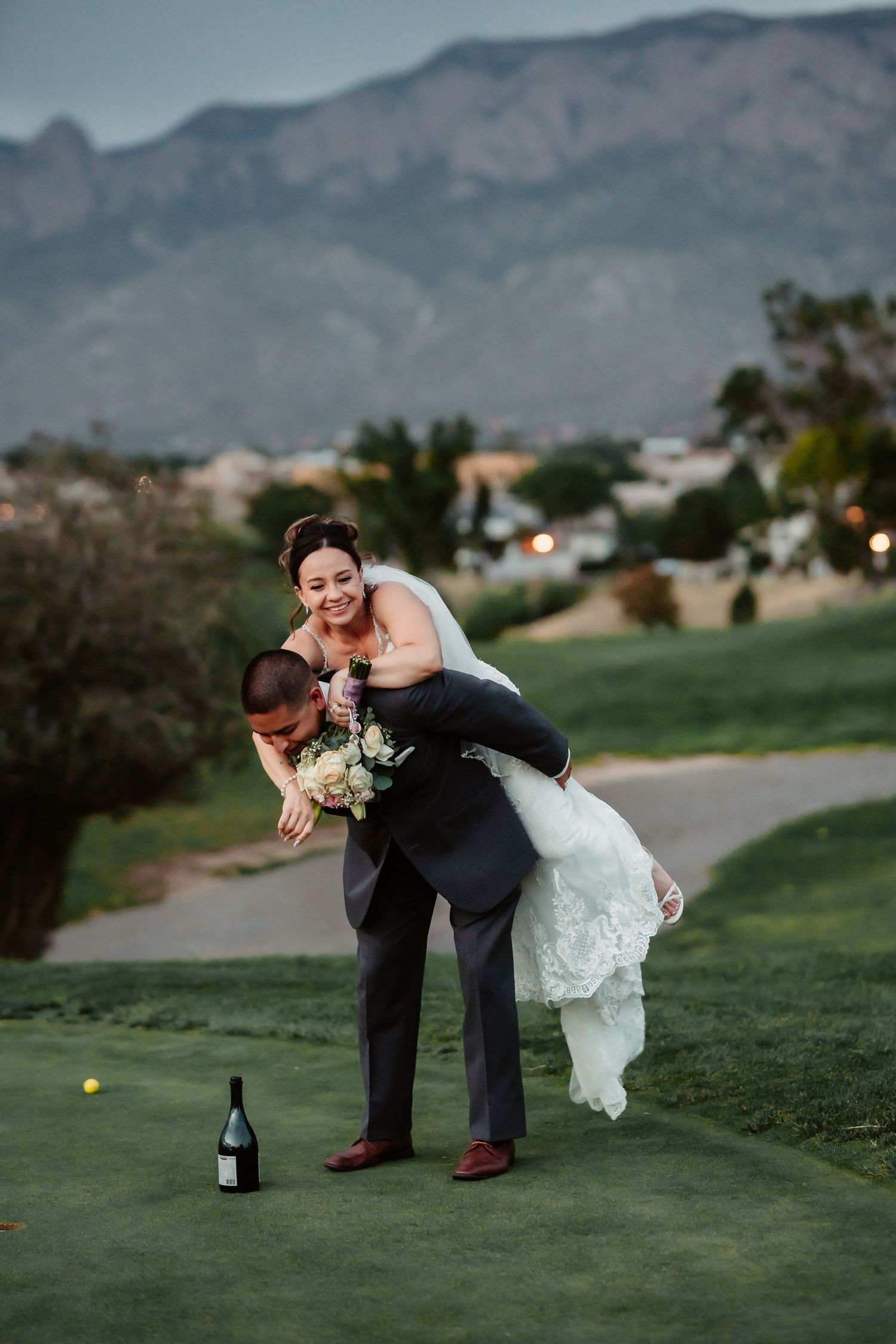 A playful newlywed couple poses on a golf course with scenic mountain views at dusk.