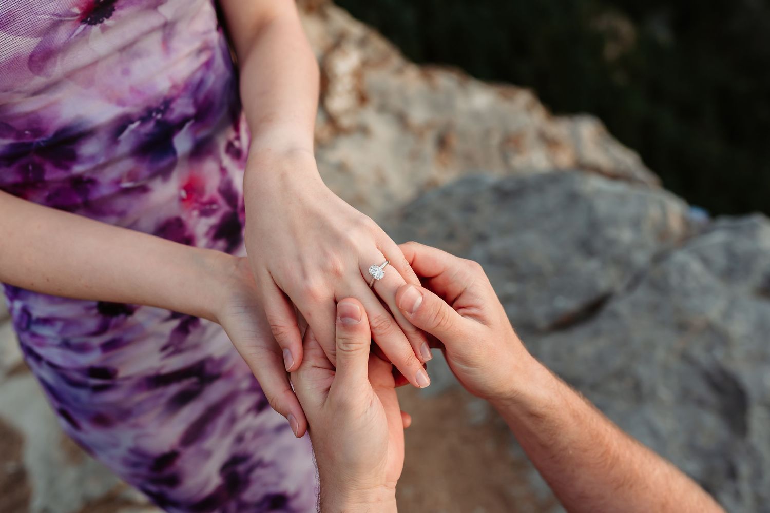 Close-up of hands exchanging an engagement ring on a rocky mountain overlook.