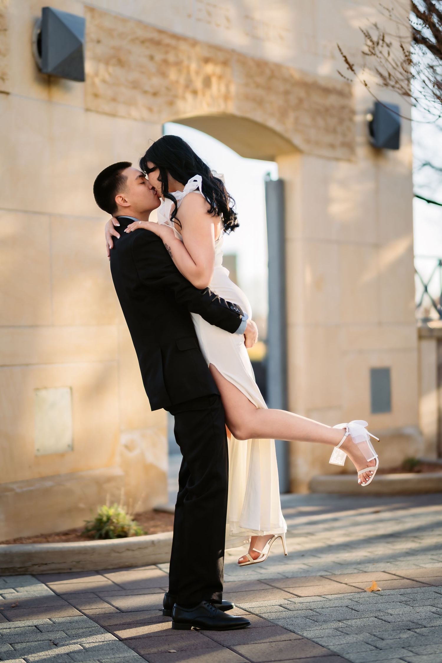 A romantic couple embraces in an outdoor courtyard setting, with the bride wearing a white dress and heels.