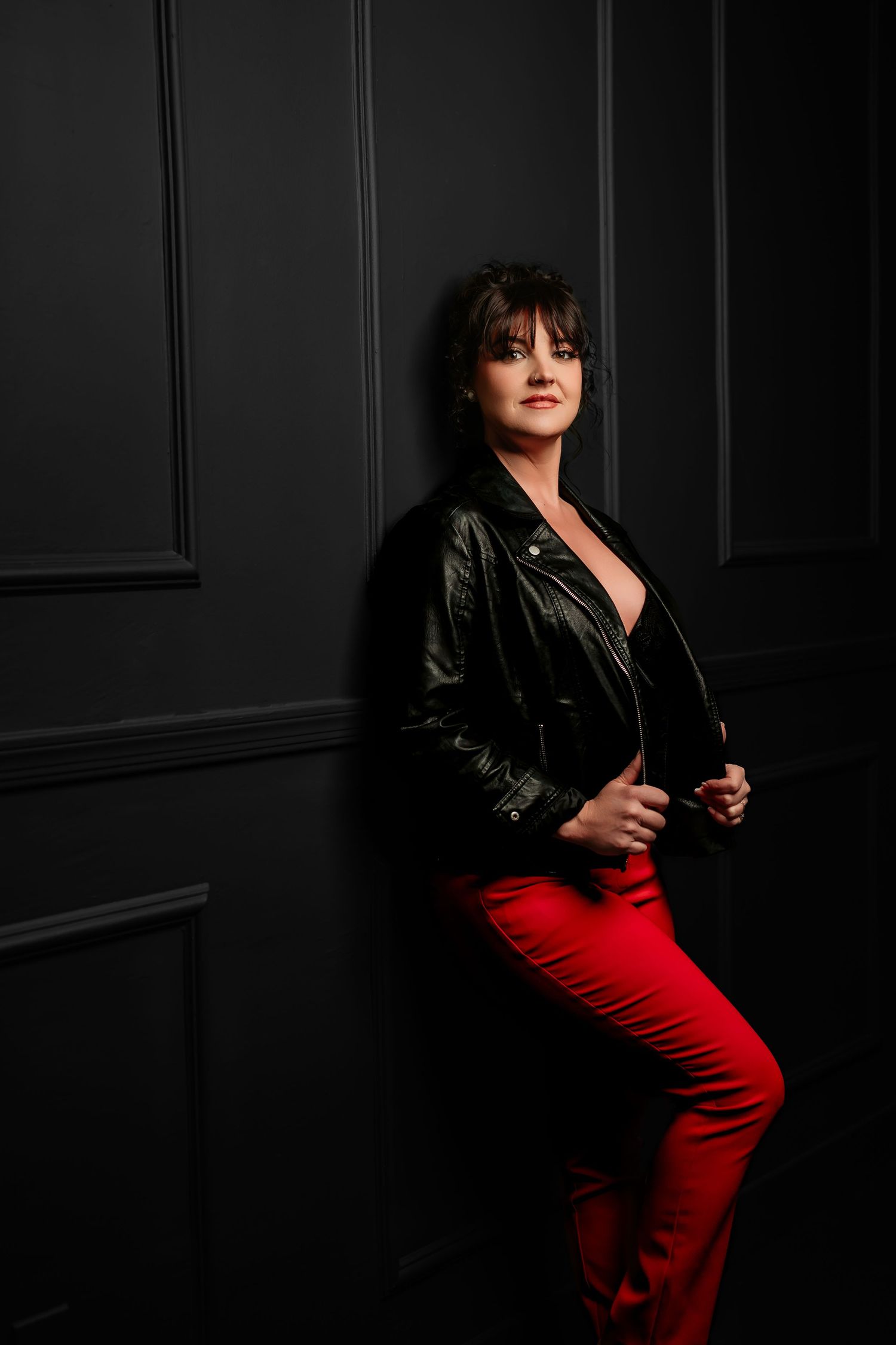 Dramatic portrait of someone wearing a black leather jacket and red pants against a dark wall with moody lighting.