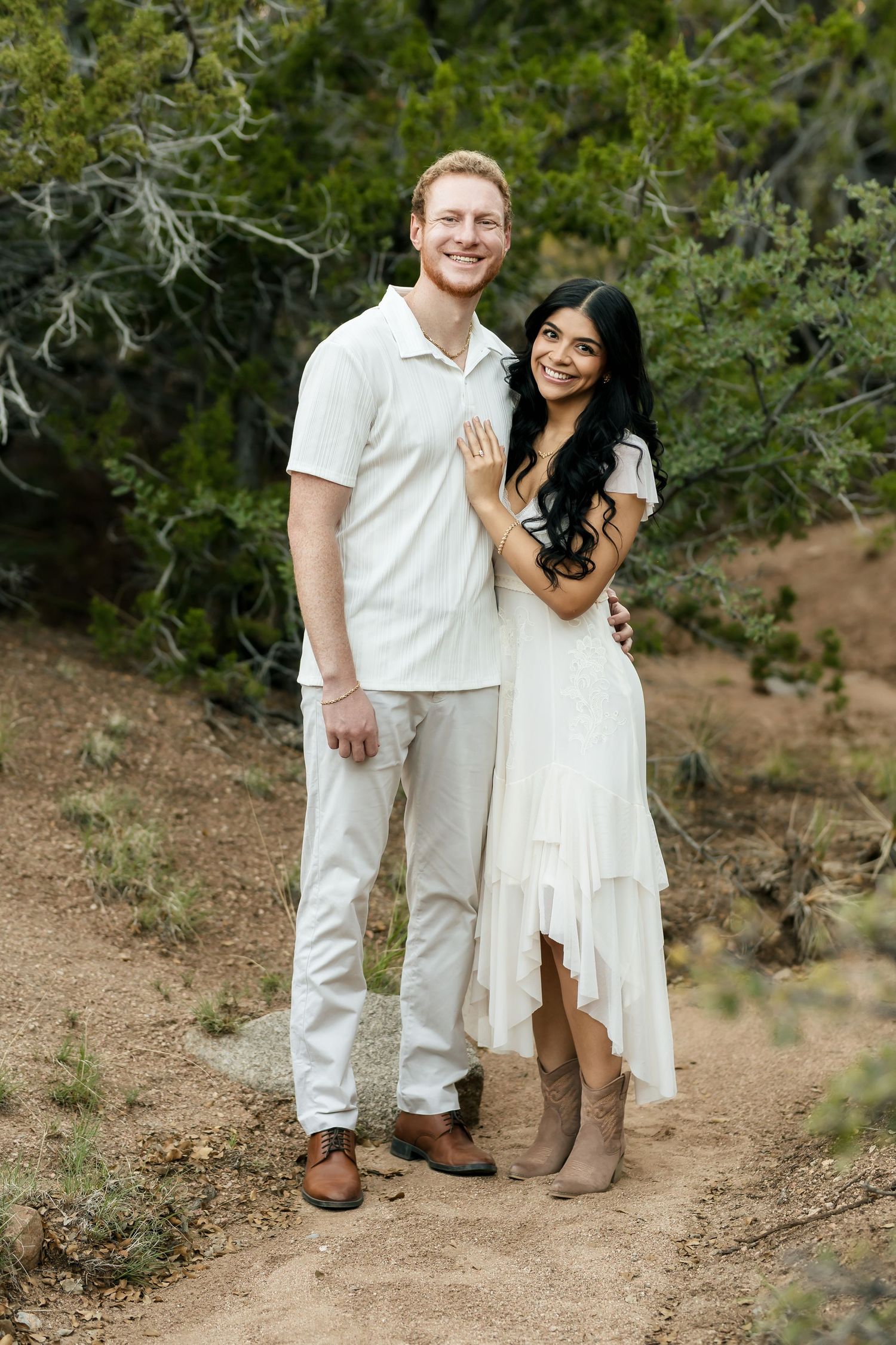 Couple in matching white outfits smiling together on a dirt path surrounded by green trees outdoors.