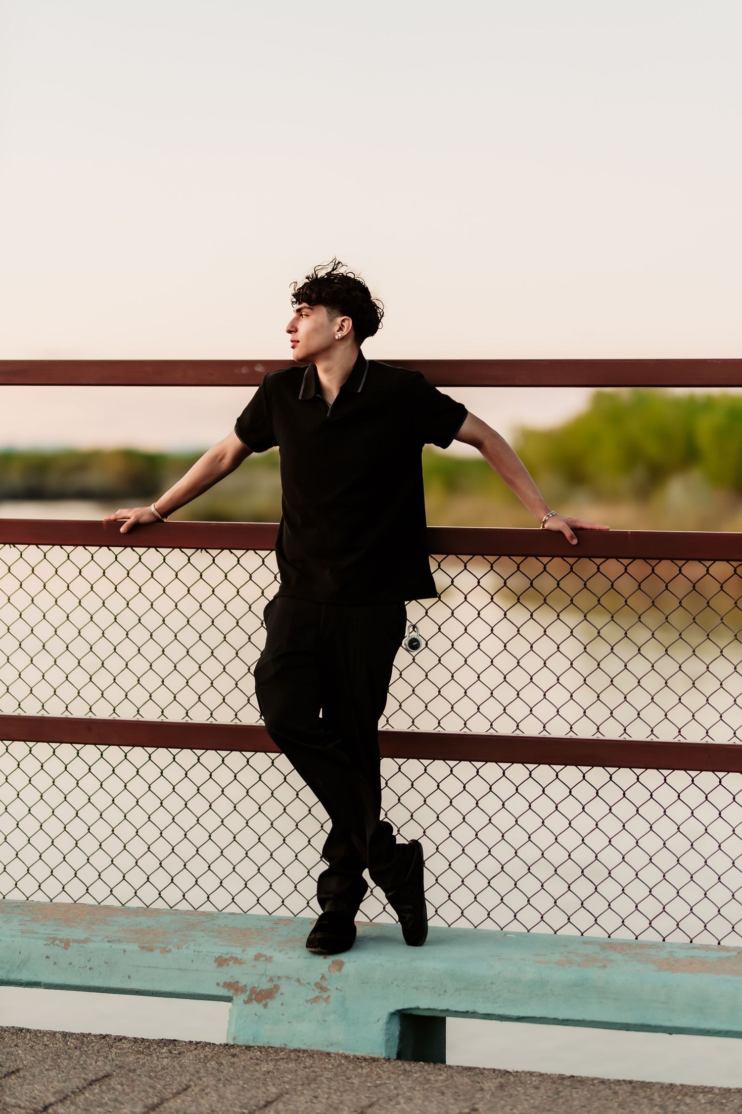 A person in all black clothing stands against a chain link fence during sunset, creating a moody silhouette.
