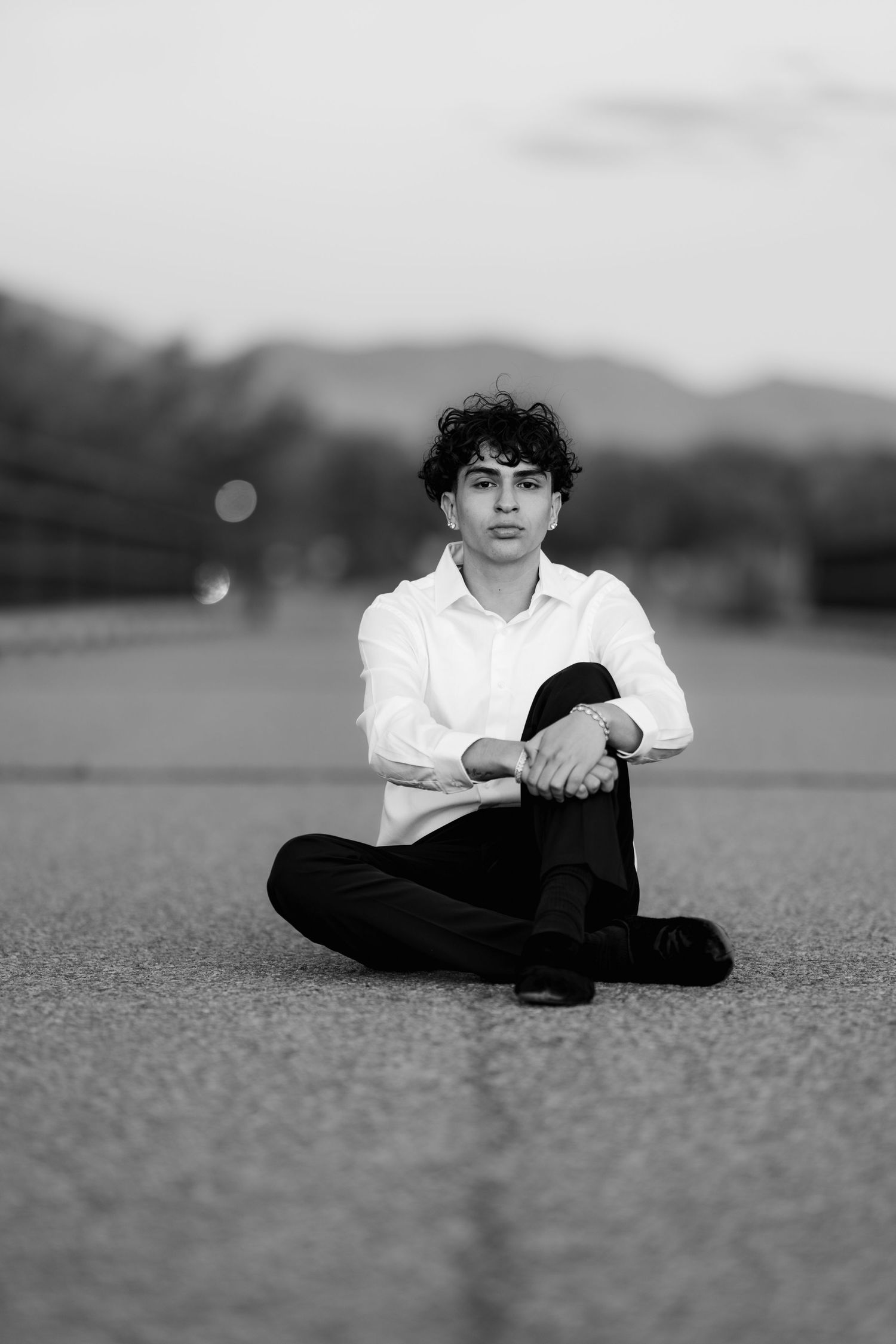 Black and white portrait of a person in a white shirt sitting cross-legged on an outdoor surface with mountains in background.