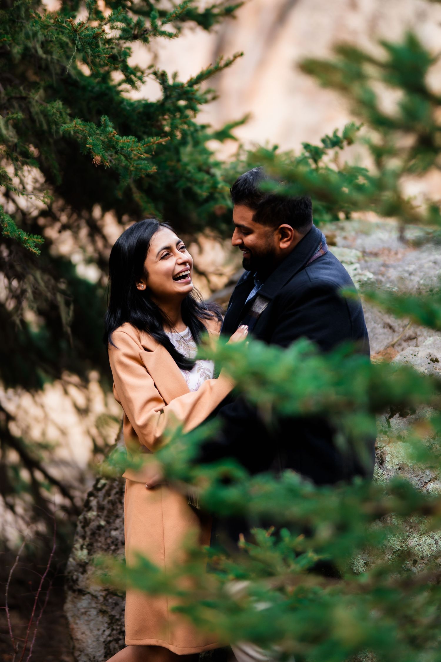 A couple laughing together among evergreen trees during an outdoor photo session.