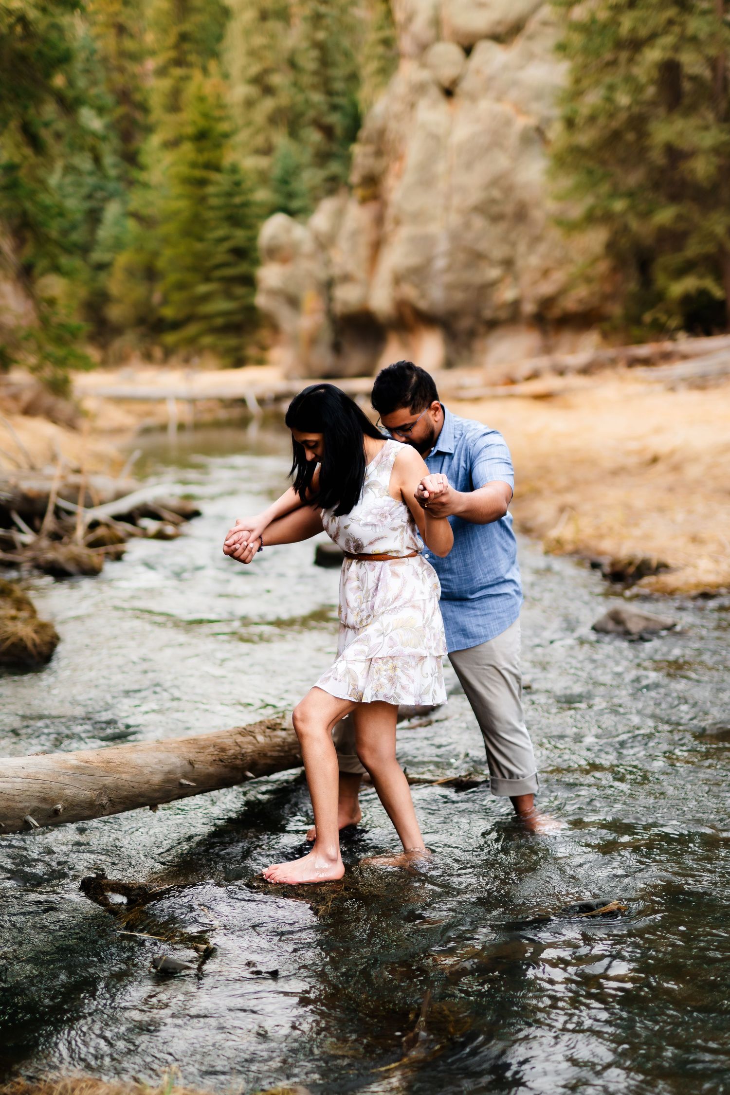 A couple carefully crosses a log over a mountain stream while holding hands.