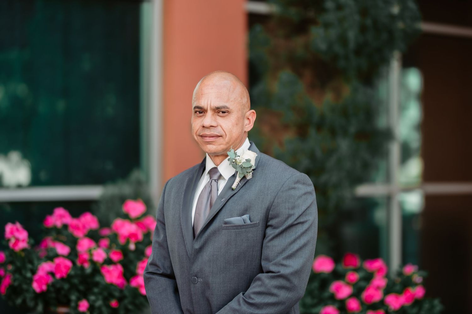 A person in a grey suit stands in front of pink flowers and bushes outside a building.