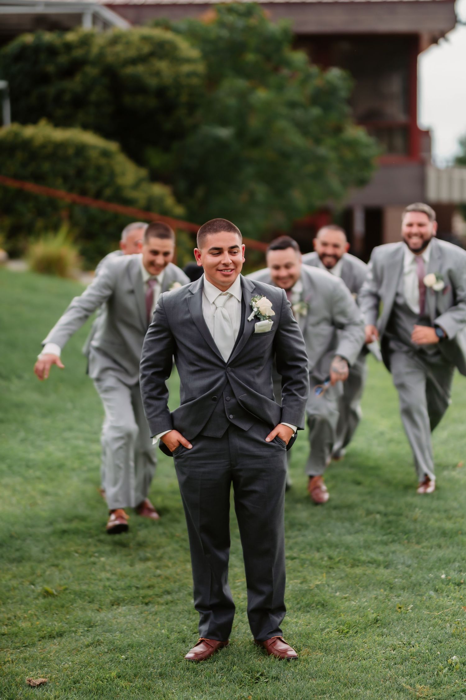 Groomsmen in matching gray suits run playfully across a grassy lawn during wedding photos.