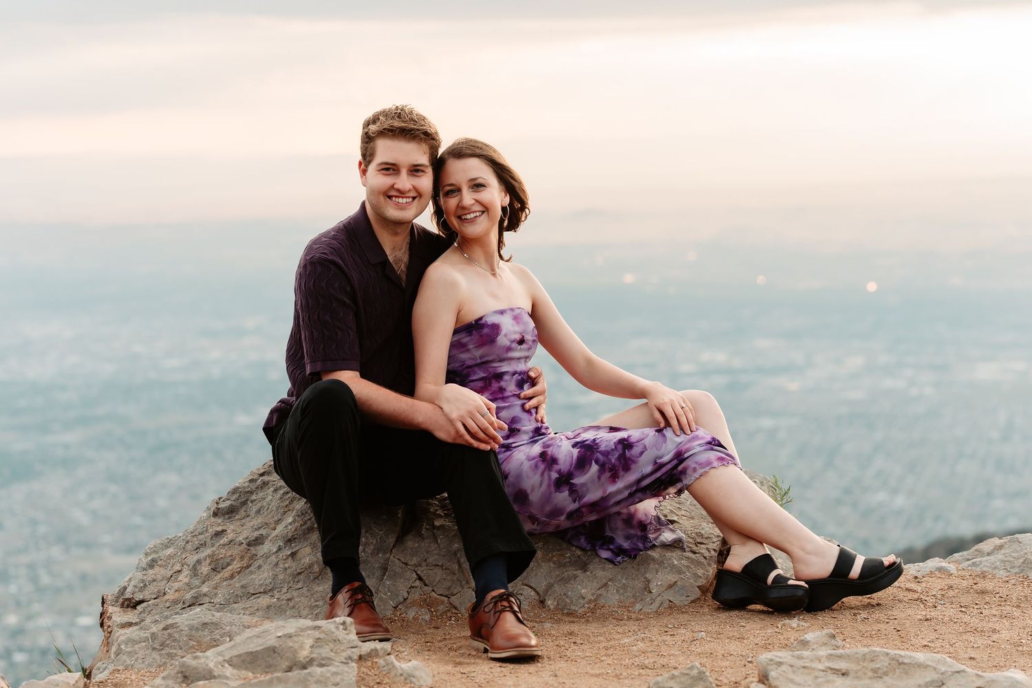 Couple embraces on rocky mountain ledge during golden hour with scenic valley vista below.